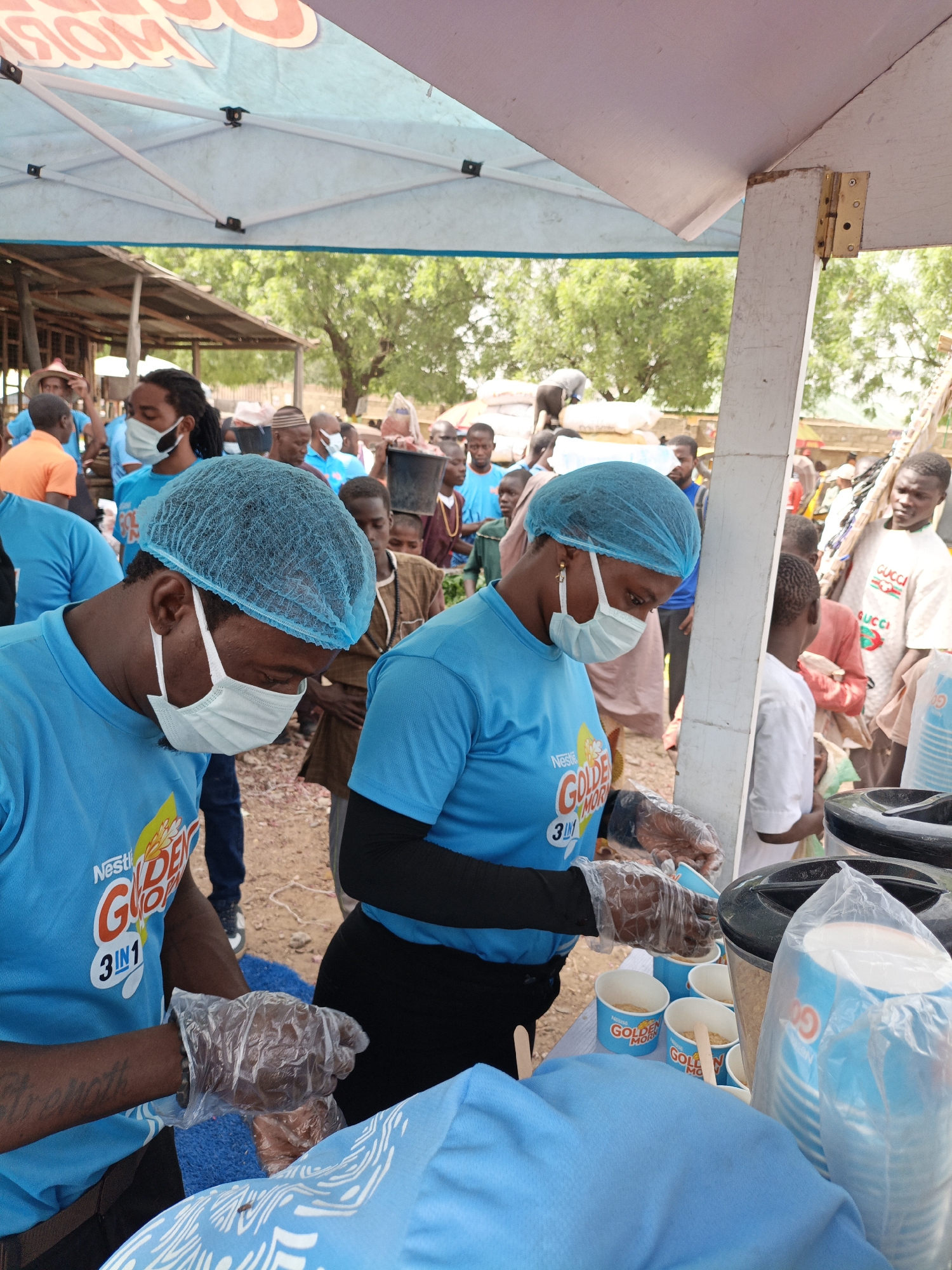 Here is a detailed description of the image:
**Scene:** The photo shows a group of people wearing blue shirts and blue hairnets, likely employees of Nestlé, distributing samples of "Golden Morn 3 in 1" cereal. The setting appears to be a market area in Kawaji, Nigeria, given the presence of other people in the background. The distribution is happening under a tent-like structure.
**People:**
* Two foreground individuals are wearing the blue Nestlé branded shirts, blue hairnets, and disposable gloves. They are likely preparing or serving the cereal samples.
* One person has a tattoo on their left forearm.
* Several other people in blue shirts and masks are in the background.
* A diverse group of people are also in the background, including children and adults, some wearing traditional clothing. They appear to be waiting or watching the distribution.
**Objects:**
* The blue Nestlé shirts have the "Golden Morn 3 in 1" logo and branding.
* Small disposable cups and wooden spoons are visible on the table, suggesting the cereal is being served in individual portions.
* Large black containers, potentially for hot water or ingredients, are present.
* A large plastic bag filled with blue paper cups.
**Actions/Interactions:**
* The individuals in the foreground are preparing the cereal samples, placing ingredients into cups.
* People in the background are observing and possibly waiting to receive samples.
**Environment:**
* The location appears to be outdoors in a busy market or public area.
* There are other buildings, stalls, and people in the background.
* The tent structure provides some shelter for the distribution.