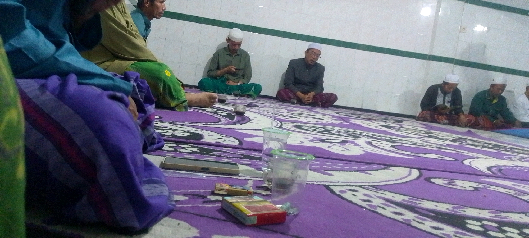 The image depicts a group of men sitting on a large, purple and white patterned carpet inside a room with white tiled walls. The men are dressed in traditional Islamic attire, including caps and long garments. Some of the men are looking at their phones, while others appear to be reading or engaged in conversation. There are also glasses of liquid and a pack of cigarettes on the carpet. The lighting suggests an indoor setting, possibly a mosque or a community gathering space. The composition of the image is from a low-angle perspective, with a portion of a person's purple and blue garment in the foreground, obscuring the lower left corner.