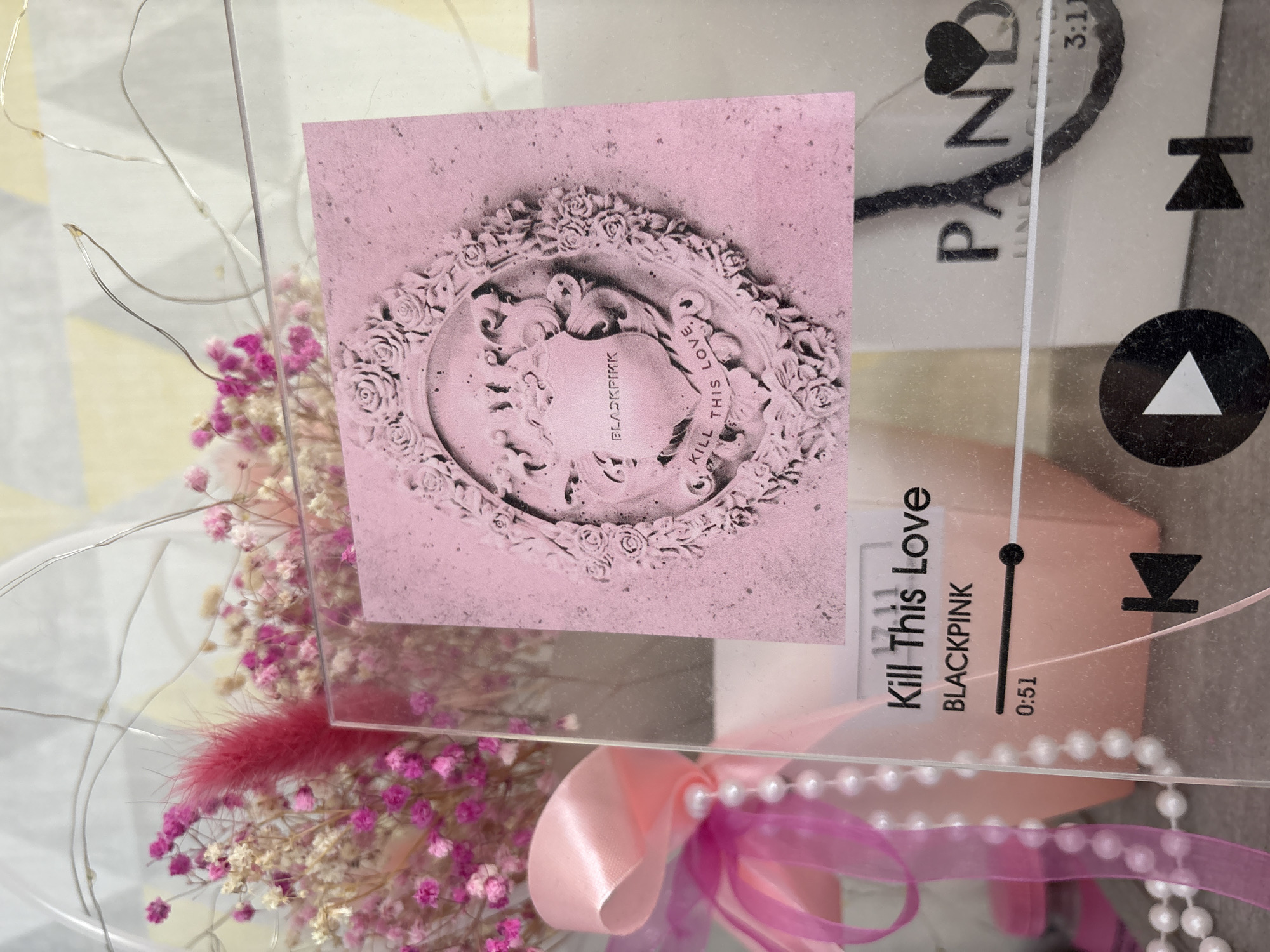 The image shows a gift arrangement. The foreground features a clear acrylic sheet printed with design elements. On the left, there's an image of the album cover for "Kill This Love" by BLACKPINK, depicted in shades of pink and grey. To the right of this, the acrylic displays a design mimicking a music player interface, also in pink and grey, with "Kill This Love" and "BLACKPINK" labeled, along with timestamps and playback controls.
Behind the acrylic, a bouquet of small pink and white flowers is visible, along with a pink ribbon and a string of imitation pearls. The background appears to be a light-colored surface.
The location of Прикамский, Russia is not discernible from this image.