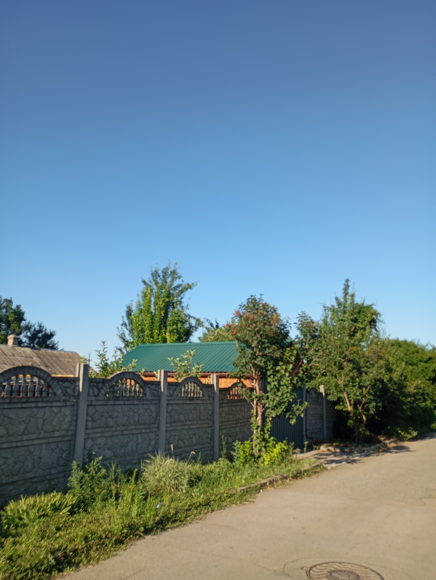 A low-angle, full shot captures a residential scene in Sozonivka, Ukraine, under a clear, bright blue sky. A segmented, grey concrete fence with decorative arched sections runs horizontally across the lower portion of the frame, separating the viewer from a property beyond. Behind the fence, glimpses of buildings are visible, including a house with a grey, tiled roof on the left and another with a vibrant green metal roof in the center. Lush green trees and vegetation surround the structures. A paved road runs along the right side of the fence, leading into the distance. The road surface shows a manhole cover in the lower right corner. The overall impression is of a quiet, rural residential area bathed in sunlight.