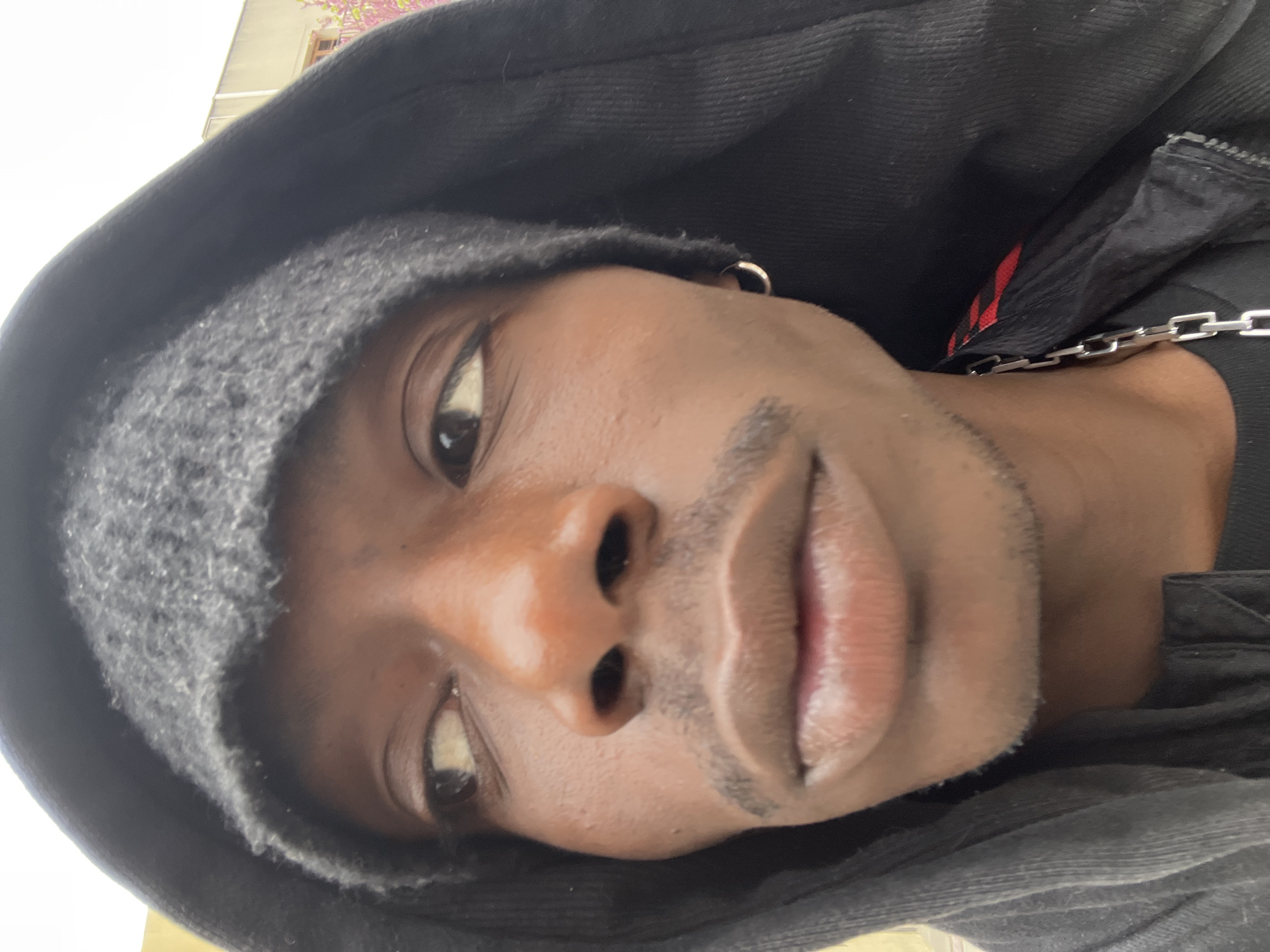 A close-up selfie shot features a dark-skinned person with brown eyes, wearing a knitted cap and a black hooded jacket. The person has sparse facial hair on the upper lip and chin. A silver hoop earring is visible in the left ear. A silver chain is draped across the person's neck and onto the jacket. Red accents can be seen on the jacket. The background is out of focus, preventing identification of the location. There is no indication that the photo was taken in Barcelona, Spain.