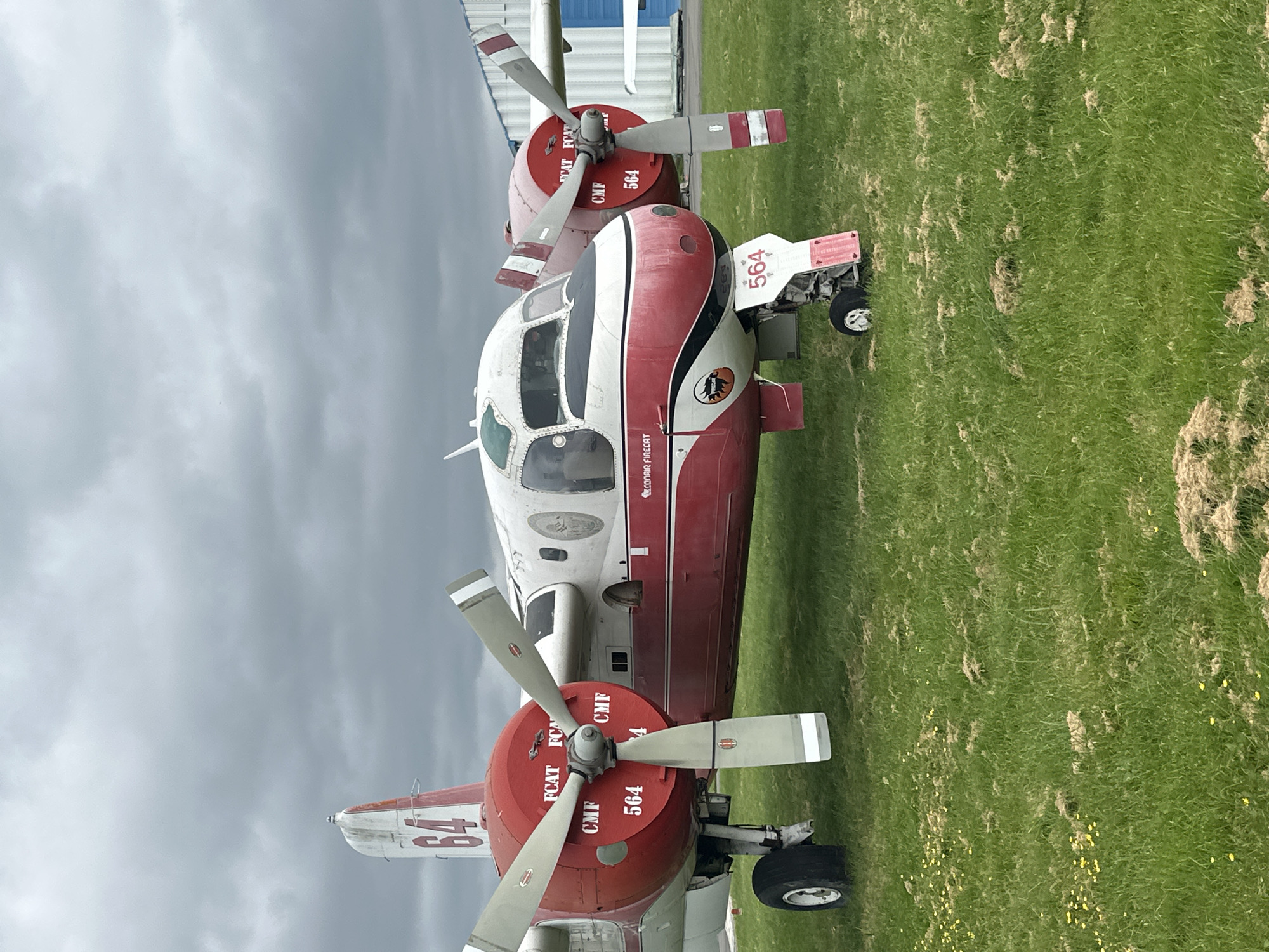 The image depicts a red and white Conair Firecat aircraft, number 564, parked on a grassy field under an overcast sky. The aircraft has two propellers and is equipped with landing gear. The location is the Township of Langley, Canada, based on the aircraft's markings indicating Conair, a Canadian company based in Abbotsford. There are no people visible in the image. The environment consists of grass and a partially visible building.
