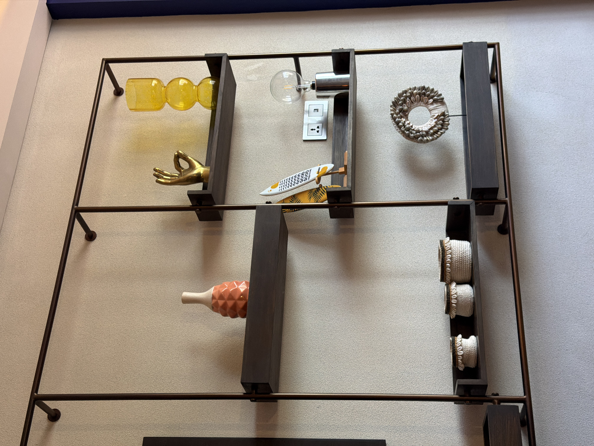 A decorative metal shelf hangs on a wall. The shelf is composed of a dark brown metal frame holding several darker brown rectangular wood shelves. On the top row from left to right are an orange glass bar, a gold-colored hand sculpture, a light bulb on a metal base, an electrical outlet, a small surfboard on a wood stand, and a round wreath-like object. The second row features an orange pineapple-shaped object and a set of three decorative white containers with brown accents. The photo may have been taken in Kuta, Indonesia.