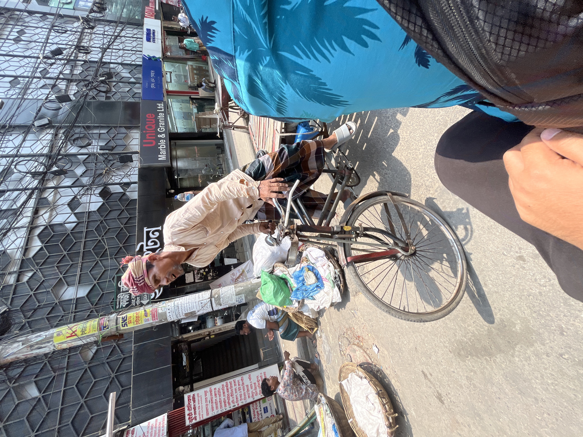 The image shows a street scene in Dhaka, Bangladesh. In the foreground, an older man wearing a light-colored shirt and checkered lungi is riding a bicycle loaded with textiles and other materials. The cyclist is positioned slightly to the left of center. A person partially obscures the upper right of the frame.
Behind the cyclist, several people are standing or sitting on the sidewalk near shopfronts. To the left, a multistory building has a geometric facade and is adorned with various signs and advertisements in Bengali and English, including a sign for "Unique Marble & Granite Ltd." Wires and cables are tangled along the building's facade. The street is paved and appears to be in daylight with sunlight on the right.