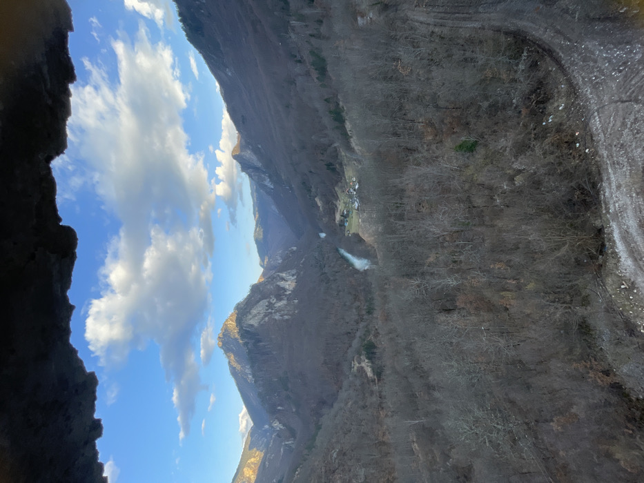 The image presents an elevated, panoramic view of the Tara River Canyon in Đurđevića Tara, Montenegro. The immediate foreground is a dark, irregular rock overhang spanning the top of the frame. Below this, a vast valley unfolds under a blue sky adorned with scattered white cumulus clouds.
Steep, rugged mountains, primarily covered with bare deciduous trees, dominate the mid-ground and background, suggesting a late autumn or winter season. A light blue-green river, the Tara River, snakes through the deepest part of the valley floor. Adjacent to the river, a small cluster of buildings with visible roofs indicates a localized settlement. Peaks in the distance feature rocky exposures. The lower right section of the image reveals a dirt road or track with visible tire impressions and scattered debris, bordering a forested incline. No individuals, vehicles, or dynamic actions are present in the scene.
