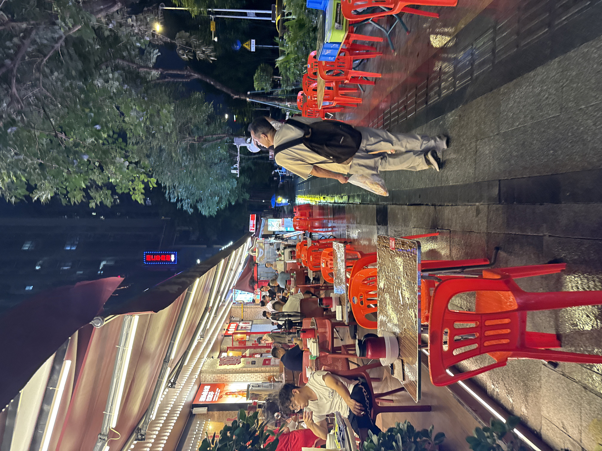 The image depicts an outdoor seating area of a restaurant at night in Haizhu District, China. The scene is lit by the restaurant's awnings and a street lamp. Red plastic chairs and tables are set up along a wet sidewalk, indicating it may have recently rained. Several patrons are seated at tables, some eating and drinking. In the foreground, a man with glasses is seated at a table, holding a cup to his lips. Further back, other diners and staff are visible. To the right of the frame, a man wearing a backpack walks away from the camera, holding a plastic bag. The background shows a building with illuminated windows and trees lining the street.