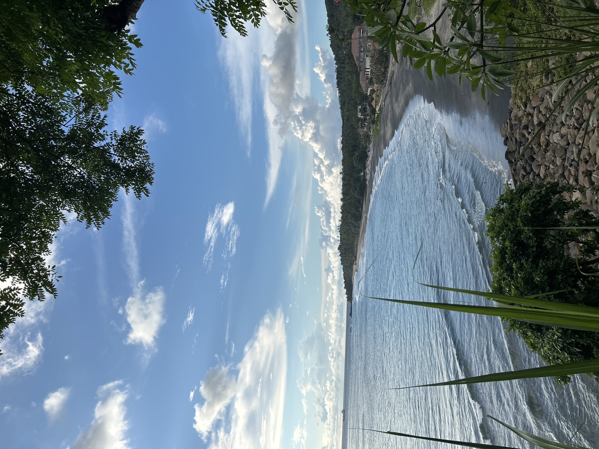 The photograph captures a coastal scene in Senggigi, Indonesia. The view is framed by foliage in the foreground, specifically trees on the left and shrubs/grasses on the right. A black sandy beach stretches along the coastline, bordered by a line of trees. The ocean, with visible wave patterns, occupies a significant portion of the image. In the distance, a building or structures are visible along the coast. The sky is blue with scattered white clouds.