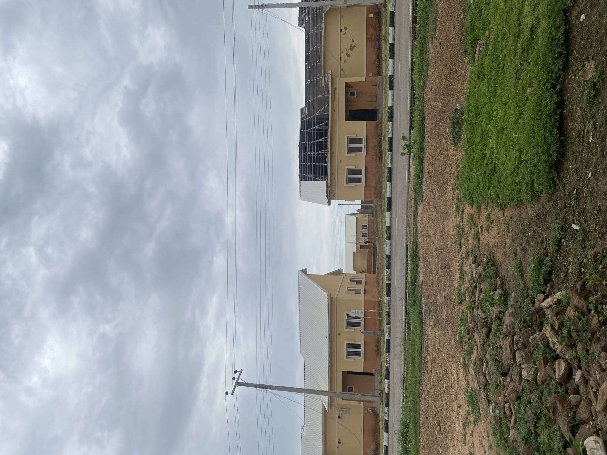 This media file captures a street view of an ongoing construction project in Bulamare, Nigeria. The scene is dominated by two multi-story buildings, one of which appears to be in a more advanced stage of construction, with its roof structure exposed. The buildings have a beige exterior with some areas showing signs of weathering or unfinished plasterwork. They are lined up along a paved road marked with black and white borders.
To the right of the road, there is an area of overgrown green grass and a patch of earth with scattered rocks and small plants. To the left of the buildings, a power pole with electrical wires stretches across the frame, indicating the presence of utilities. The sky is overcast with heavy gray clouds, suggesting a cloudy or potentially rainy day. The overall lighting is diffused, and the time of day is difficult to ascertain due to the weather conditions. There is no discernible text visible in the image. The scene evokes a sense of ongoing development and the typical urban landscape of a developing area.