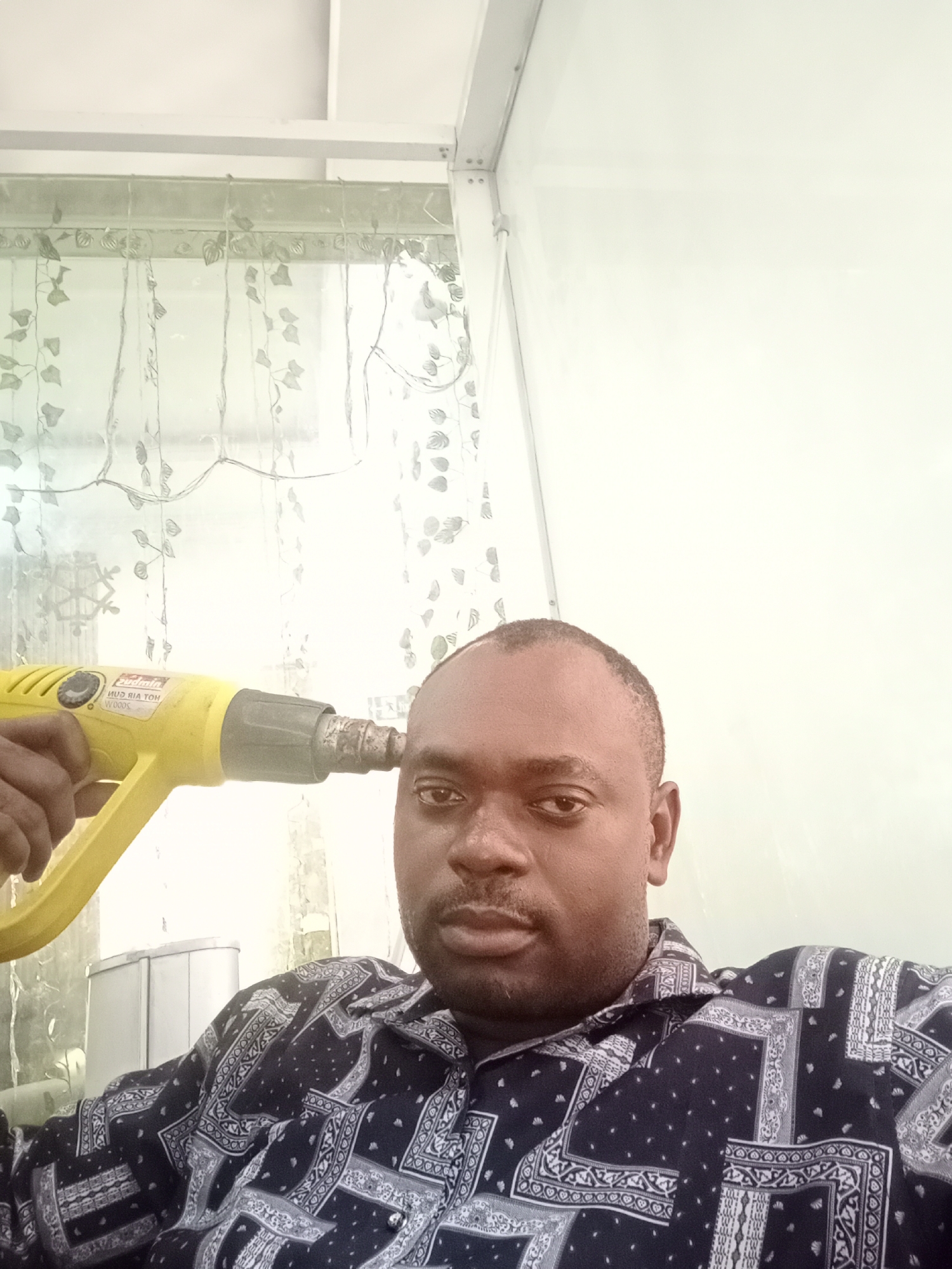 A man is holding a yellow heat gun to his head. He is wearing a patterned dark blue shirt. A window with sheer, vine-patterned curtains is visible in the background. A white wall and ceiling are also visible. The location is Gwarinpa, Nigeria.