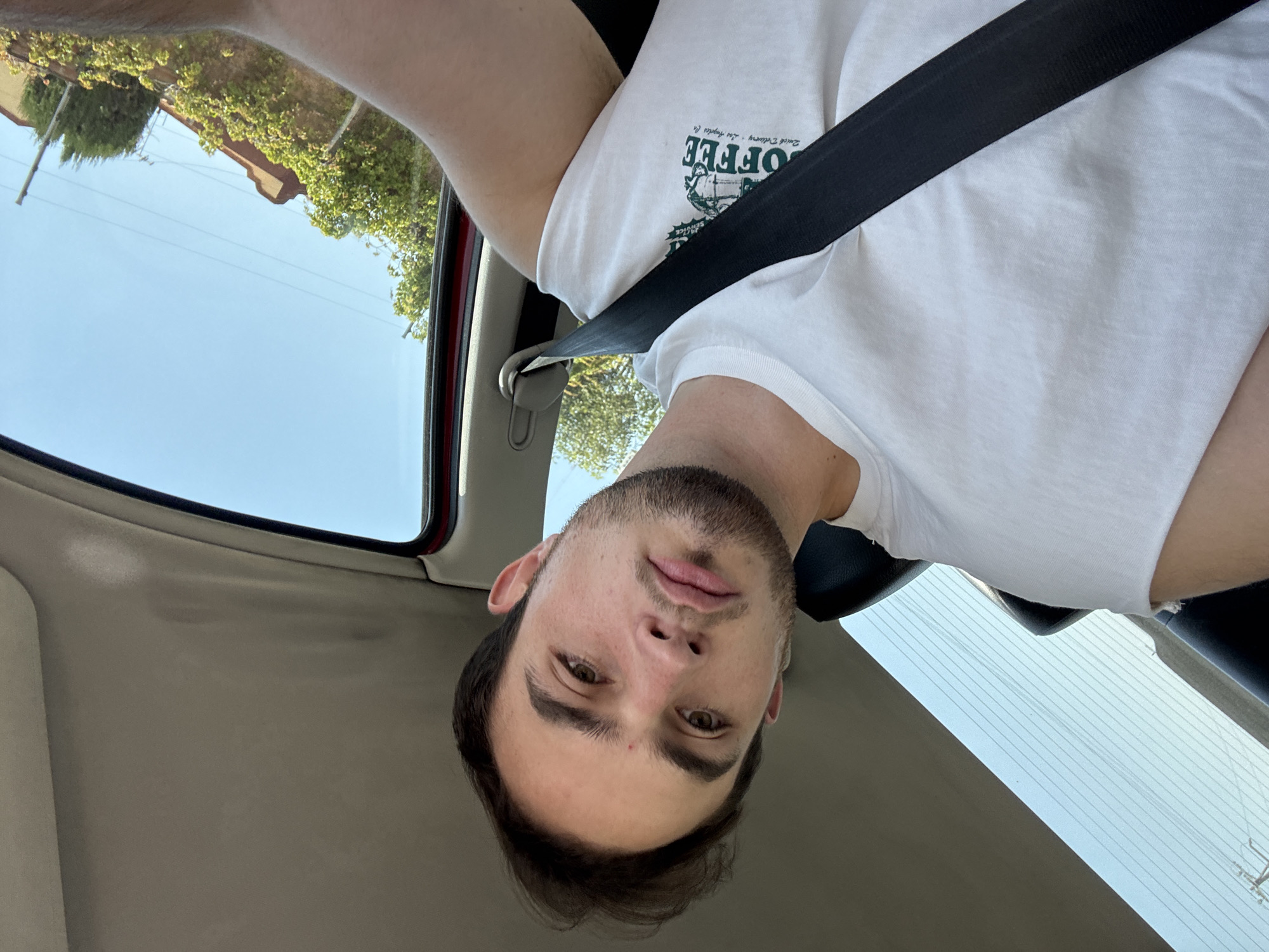 A young man is in a car wearing a white t-shirt and a seatbelt. The car is likely in San Bernardino Tlaxcalancingo, Mexico. The view out the window shows a blue sky, power lines, and a few buildings.