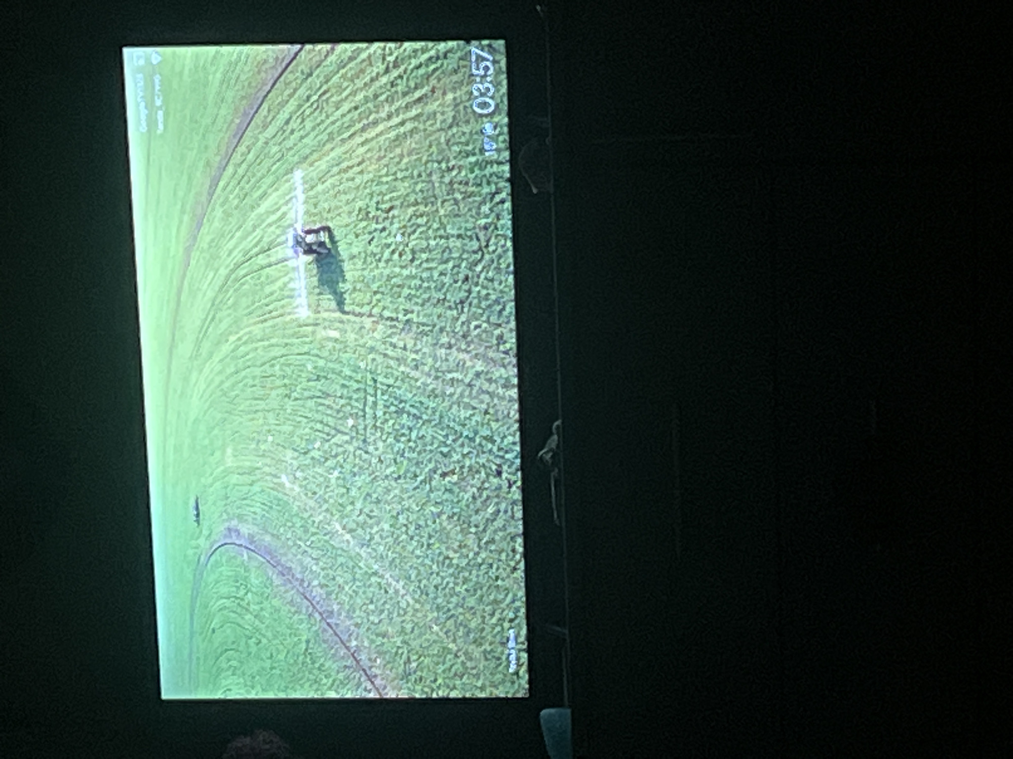 The image displays a projection screen showing a drone view of a vast green field with circular irrigation patterns. In the center of the frame, a small agricultural drone is visible, actively spraying the field. The lower right corner of the screen shows a timestamp indicating 03:57 and a temperature of 15 degrees. The projection screen is set up in a dark room, with the lower portion of the image showing the top of a dark piece of furniture. The overall scene depicts an agricultural operation likely taking place in the city of Zarudyntsi, Ukraine.