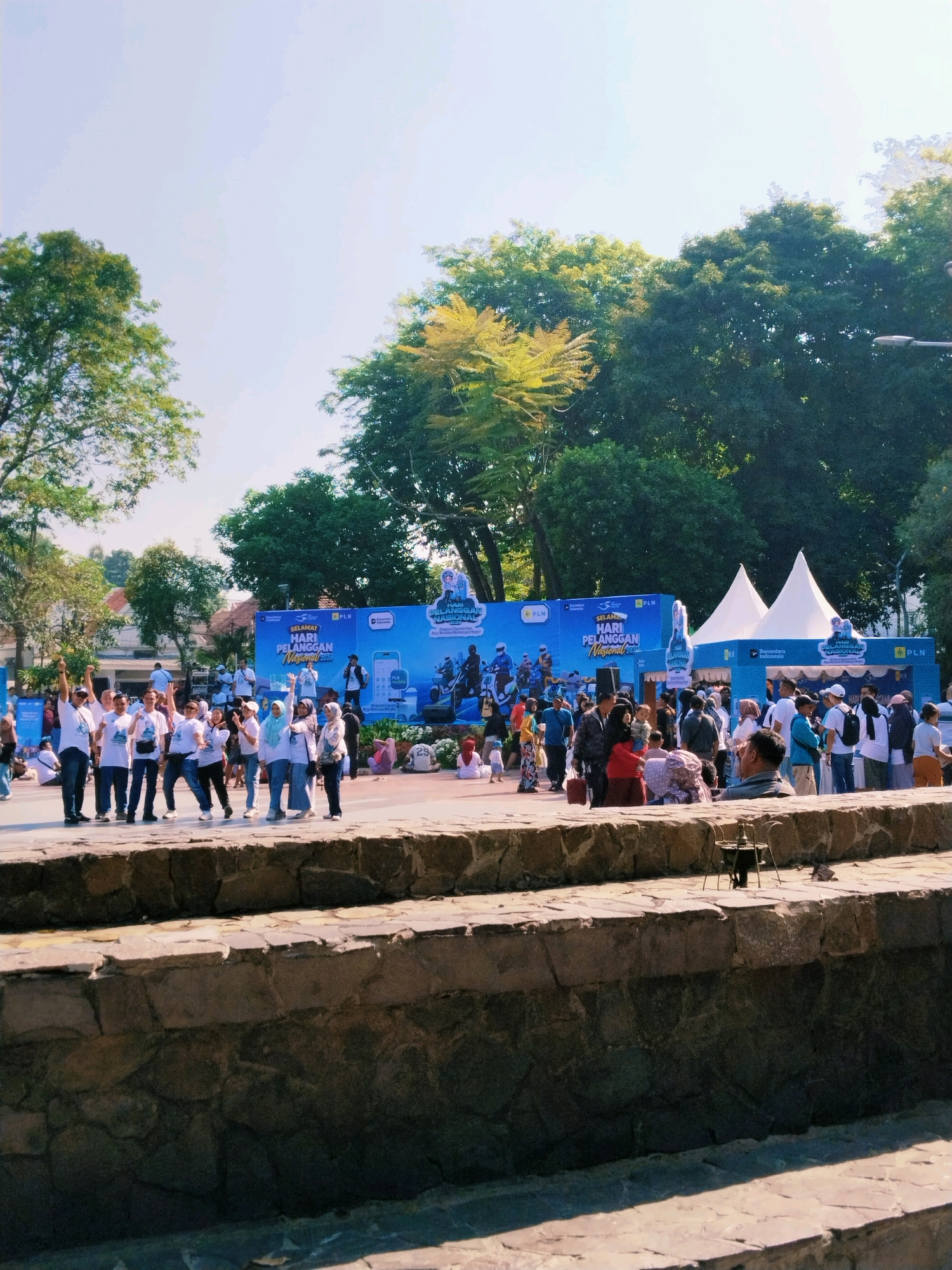 The image depicts an outdoor event in front of a large blue banner with text that appears to be in Indonesian. The banner has images of people on motorcycles and what look like electric scooters. There are two white tents set up behind the banner. A group of people, mostly dressed in white t-shirts, are posing for a photo in the foreground. They are standing on a paved area. Behind them, a crowd of people are gathered, some interacting with booths and tents. In the background, there are trees and buildings. The overall atmosphere suggests a public event or festival. The text on the banner mentions "SELAMAT HARI PELANGGAN NASIONAL 2023" which translates to "Happy National Customer Day 2023". Given the context of National Customer Day and the PLN logo (which represents PT PLN (Persero), the state electricity company of Indonesia), it is highly likely this event is taking place in Indonesia, possibly in Surabaya, as indicated by the provided context. The stone structure in the foreground suggests it might be a park or public space.