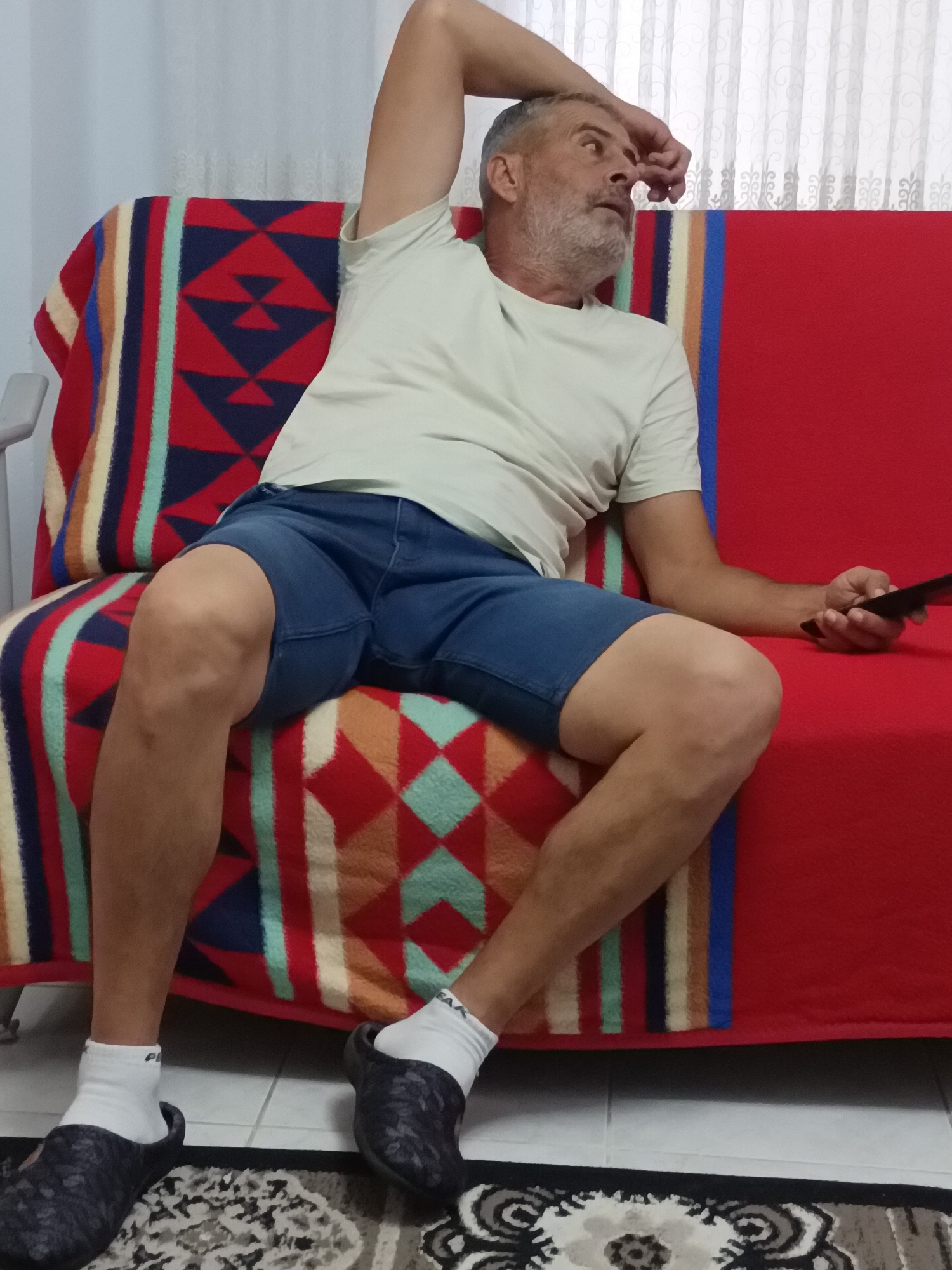 A middle-aged man with graying hair and a beard sits on a red sofa covered with a patterned blanket. He is wearing a light-colored t-shirt, blue shorts, white socks, and dark clogs. His right arm is raised, with his hand resting on his forehead, and he is looking to the right. In his right hand, he holds a black object, likely a remote control. The sofa is positioned in front of a white curtain. The flooring appears to be tile with a rug in front of the sofa. The environment suggests a domestic setting, possibly a living room. The image is a full-body shot of the man and the immediate surroundings of the sofa. There is no discernible information about the location being in Alanya, Turkey.