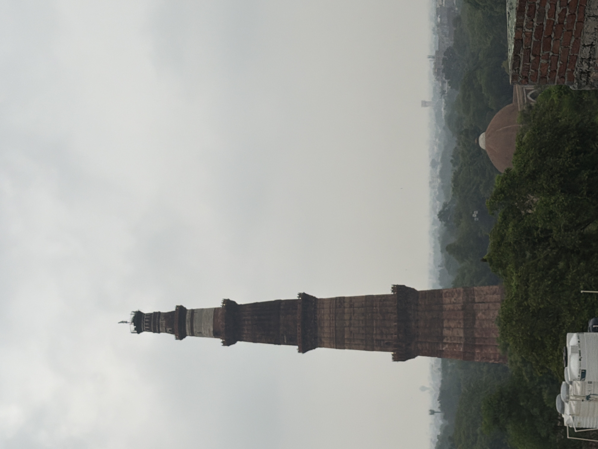 The image shows a view of the Qutub Minar in Delhi, India, under a cloudy sky. The tall, red sandstone tower dominates the left side of the frame, with its intricate architectural details visible. In the middle ground, lush green trees surround the base of the minar, creating a dense forest-like environment. To the right and further in the background, a red dome structure, likely part of a tomb or another historical building, is partially visible amidst the foliage. A distant cityscape with some structures can be seen on the horizon. In the foreground, the top of some white water tanks is visible, suggesting a residential or urban area in the immediate vicinity. The overall atmosphere is muted due to the overcast weather.