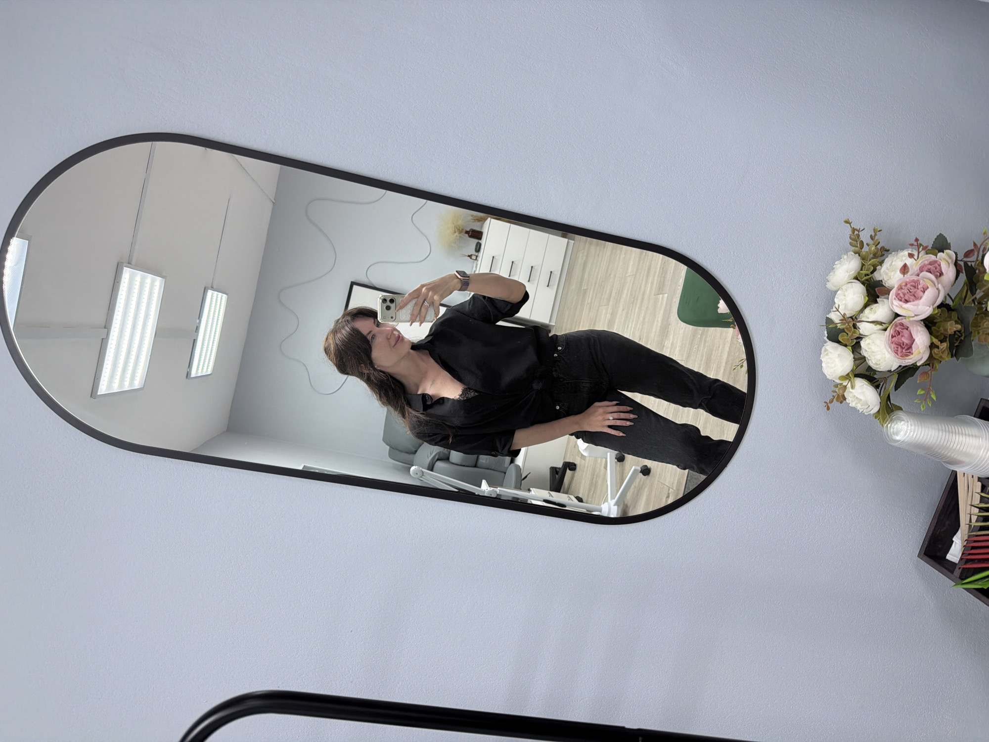 A woman takes a mirror selfie in a room with light blue walls. She is wearing a black button-down shirt with the top buttons unbuttoned, revealing a glimpse of lace lingerie, and dark wash jeans. Her hair is dark brown and styled in loose waves. She is wearing a watch on her left wrist and a ring on her right ring finger. The mirror reflects the room behind her, which appears to be a professional space, possibly a salon or treatment room, with a white dresser, a chair, and overhead fluorescent lighting. In the foreground, a bouquet of pink and white artificial flowers sits in a vase, along with a stack of clear plastic cups and a wooden organizer containing various small items. A black metal stand is visible to the left of the mirror. The room's floor is light-colored wood. The overall impression is a clean and modern environment.