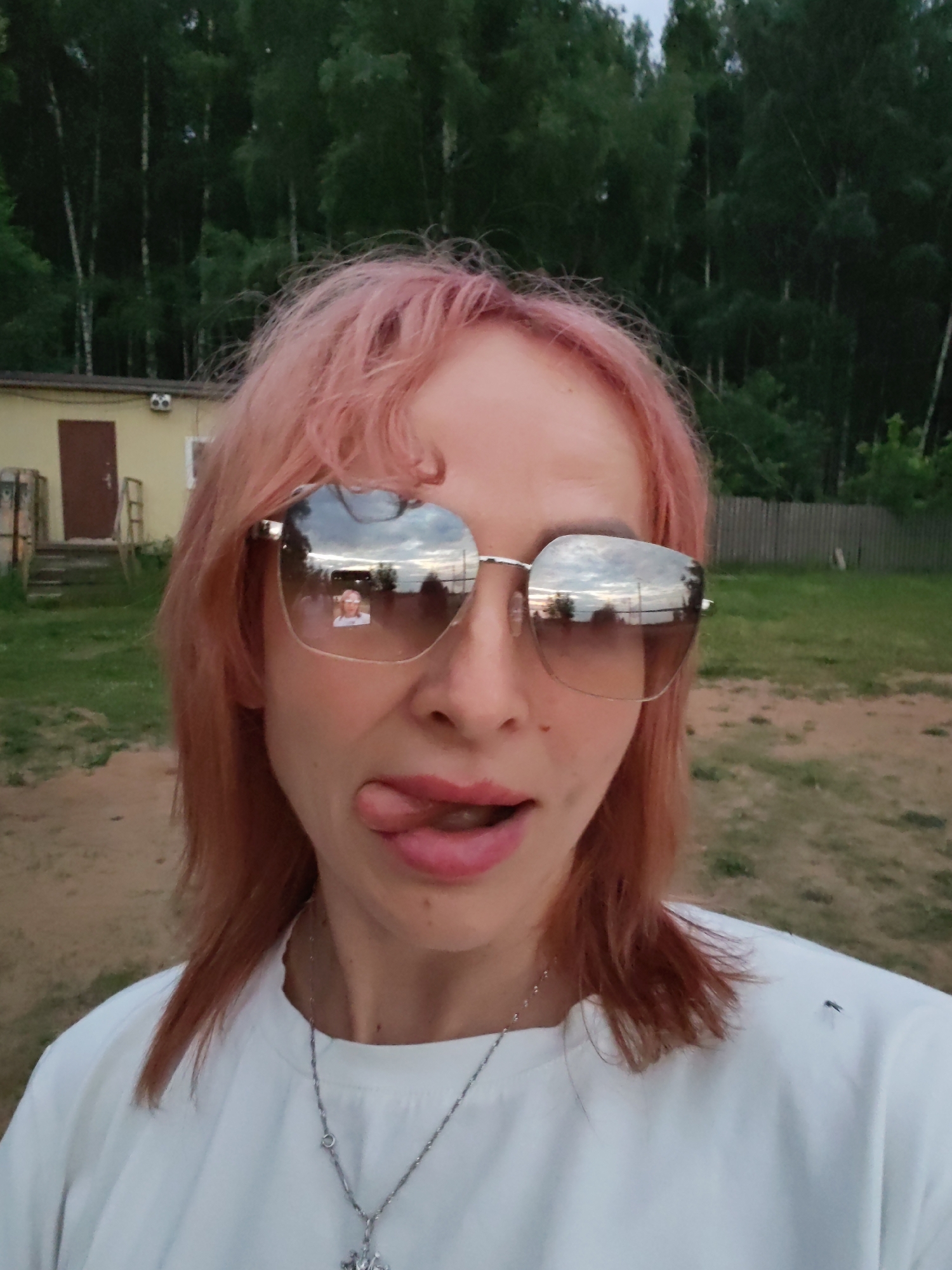 A woman with pink-tinted hair is captured in a close-up, selfie-style photograph. She is wearing large, reflective sunglasses, which show a reflection of a person and the sky. Her tongue is sticking out to the side. She is wearing a white t-shirt and a silver necklace with a pendant. The background appears to be an outdoor, rural or semi-rural setting with trees and a building that resembles a small, weathered structure. There is a wooden fence visible in the distance, and the ground is a mix of sand and grass. The lighting suggests it is either early morning or late afternoon. The location is identified as Chaikovsky, Russia.