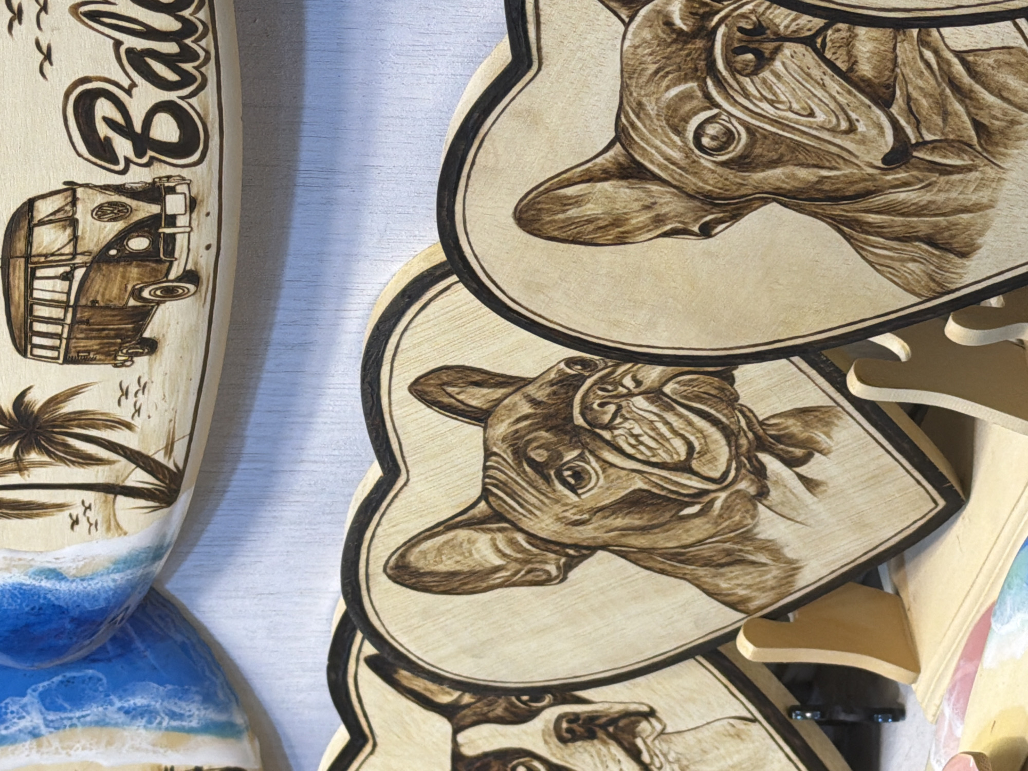 This media file features several items with wood-burned art, presented in a close-up, angled view.
The main subjects are wood-burned illustrations of French bulldogs and a vintage Volkswagen van. One wood piece is shaped like a surfboard and depicts a tropical beach scene with palm trees, birds, and crashing waves rendered in resin art. The wood-burned French bulldogs are detailed, with expressive eyes and textured fur.
The overall aesthetic suggests a collection of decorative art pieces, possibly for sale or display. The wood-burned art implies a craft or artistic creation process. The resin art on the surfboard piece adds a vibrant, glossy element.
There are no people or animals present in the scene. The setting appears to be indoors, against a lightly textured, pale background that could be a wall.
Visible text on the surfboard-shaped piece reads "Bali". The French bulldog art does not contain visible text.
The image does not provide clues about the time of day or weather. The location context of Kuta, Indonesia, might relate to the "Bali" text and the tropical imagery, suggesting a connection to surfing culture or souvenirs from that region. The emotions conveyed are through the art itself, with the bulldogs appearing somewhat playful or curious.
