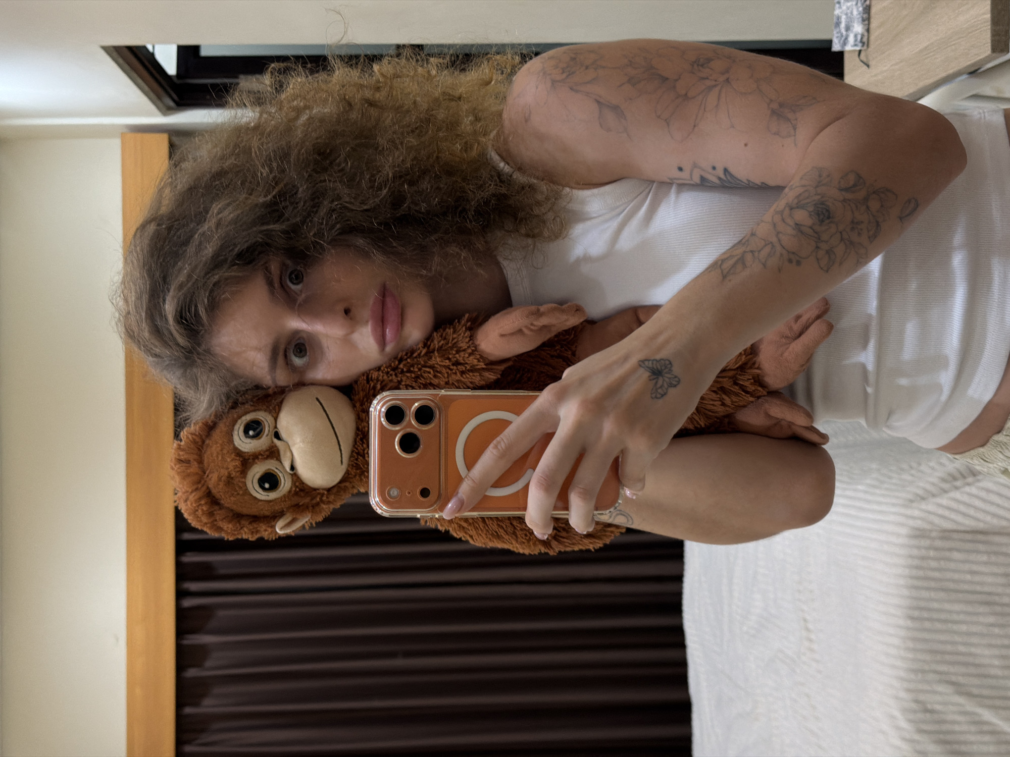 A person is taking a selfie in a bedroom in North Kuta, Indonesia, holding a stuffed orangutan. The person is wearing a white tank top and has curly brown hair. They have tattoos on their arm and a butterfly tattoo on their wrist. The stuffed orangutan is brown and fuzzy. The background of the image includes a bed with white striped bedding, a brown curtain, and a wooden headboard. A mirror is visible on the left side of the image, reflecting the scene. A phone is being held by the person's right hand to take the picture.