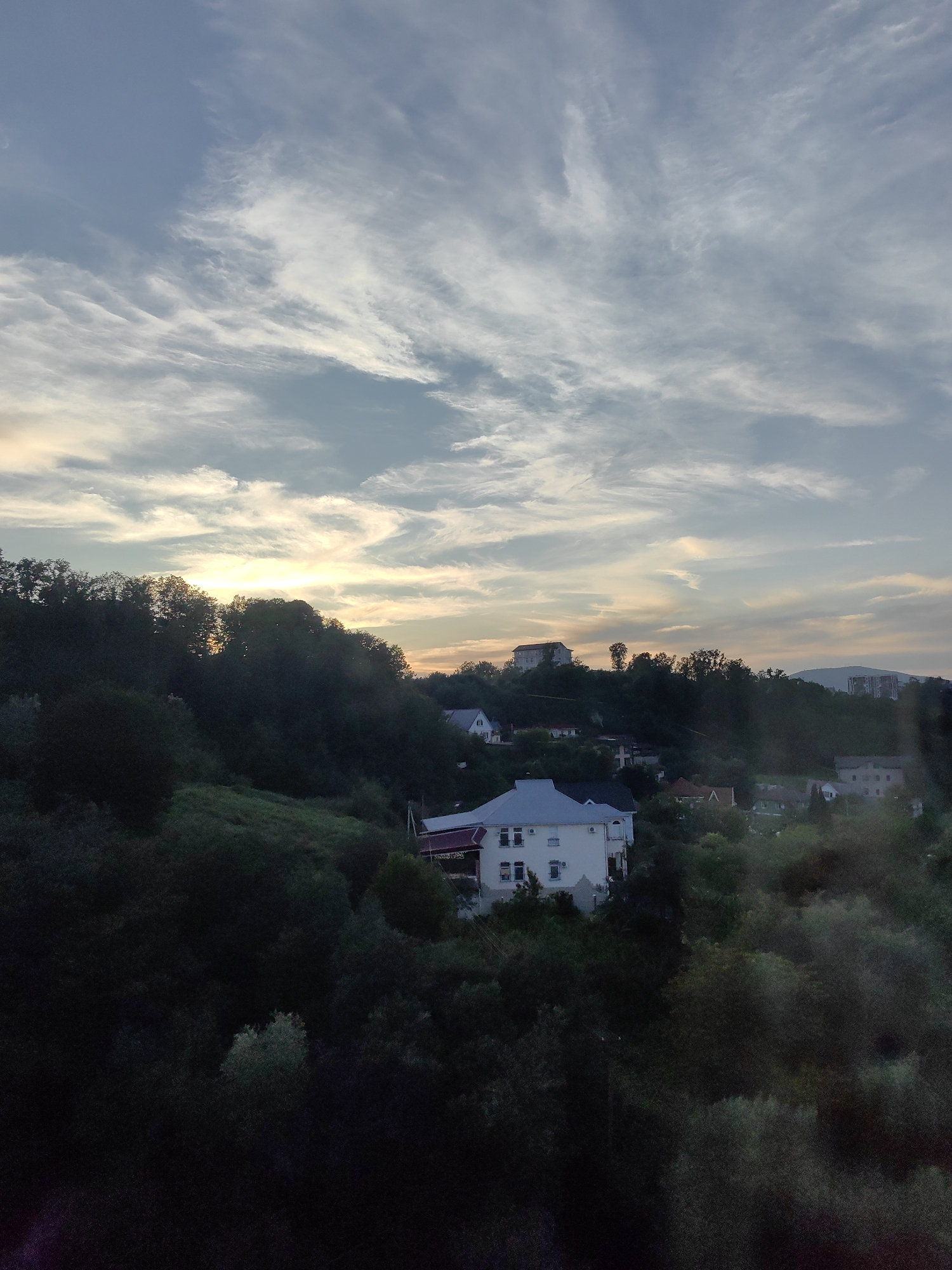 This is a medium shot, vertical, outdoor, daytime, eye-level photograph of a cloudy sky over a hillside residential area. The sky is filled with wispy, elongated clouds that are lit by the setting sun, casting a warm glow. The clouds are thin and spread out, creating a textured appearance against the blue sky.
Below the sky, a densely forested hillside slopes downwards. The trees are dark green and lush, with some lighter patches of vegetation visible in places. The dense foliage obscures much of the lower hillside.
Scattered amongst the trees, and on the slopes, are several residential buildings. In the foreground, a large, multi-story white house with a dark roof is visible. It has multiple windows and balconies. Further up the hill, other houses of varying sizes and architectural styles can be seen. Some appear to be villas or detached homes, while others might be apartment buildings. The buildings are nestled within the trees and on the slopes, indicating a suburban or semi-rural setting within a city.
The overall impression is a peaceful, scenic view of a residential area on a hillside, with the natural beauty of the trees and the dramatic sky as prominent features. The specific location is the city of Sochi, Russia, known for its subtropical climate and hilly terrain. The time of day appears to be late afternoon or early evening, given the quality of light in the sky. There are no people or vehicles visible in the image.