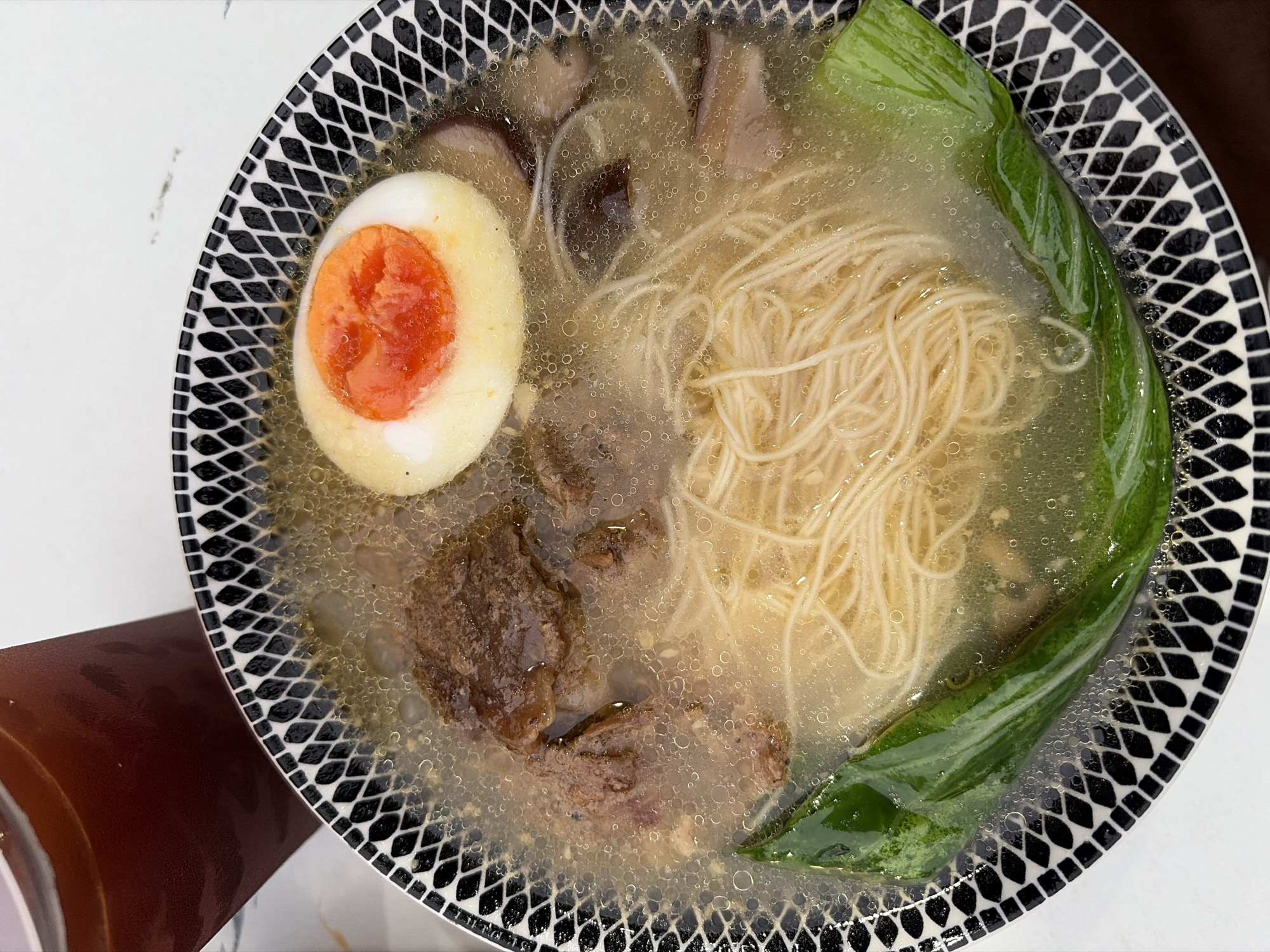 A close-up, overhead shot features a bowl of ramen with a clear broth, thin noodles, braised beef, mushrooms, a halved soft-boiled egg with a vibrant orange yolk, and a piece of green vegetable. The bowl has a black and white diamond pattern on the rim. To the upper left, a glass of dark liquid, possibly iced tea or coffee, is partially visible. The background is a white, slightly textured surface. The setting is likely a restaurant in Semarang, Indonesia, given the context.