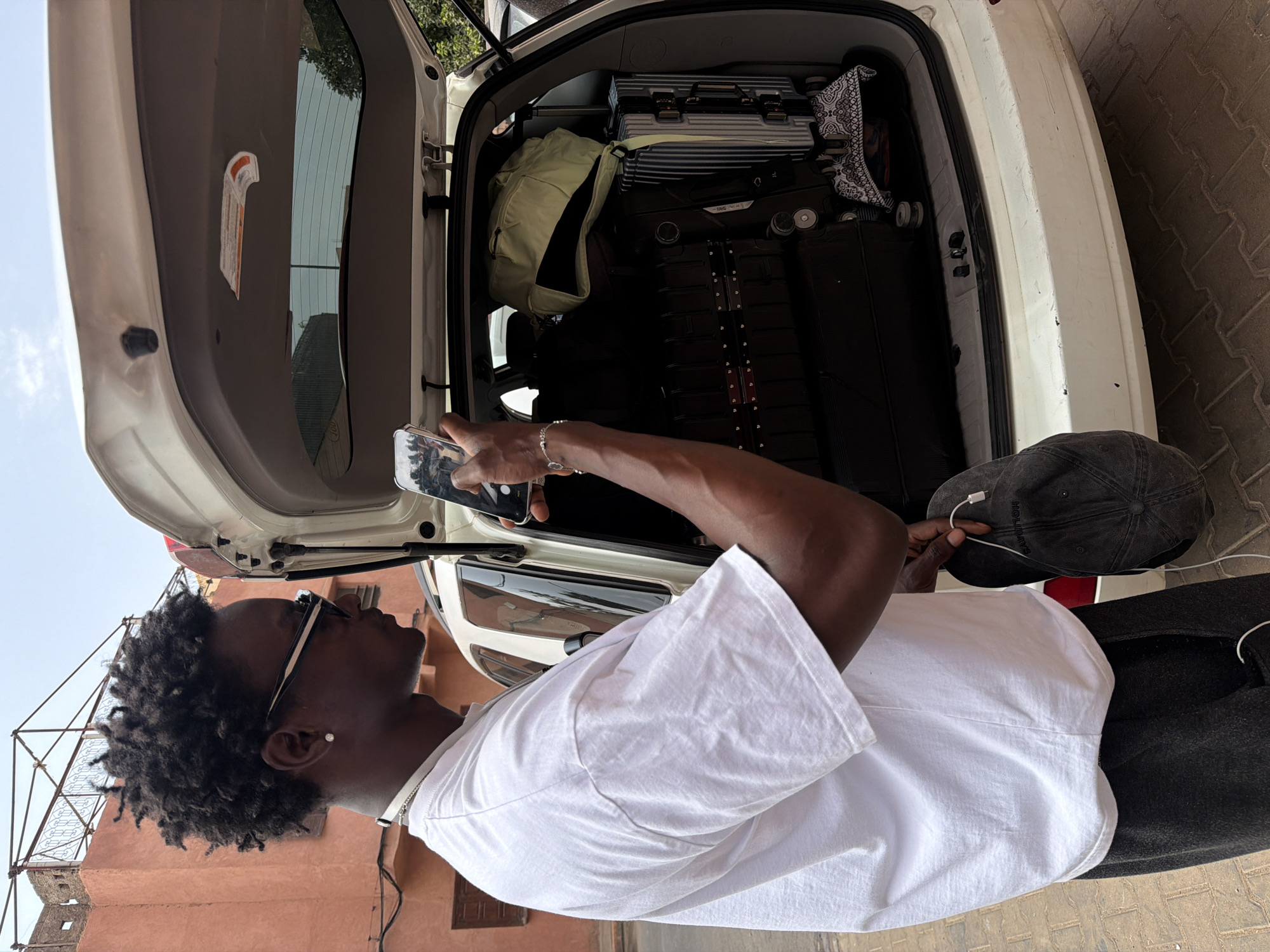 In Marrakesh, Morocco, a young Black man is standing next to the open trunk of a white SUV, holding a smartphone in his right hand, appearing to take a picture. He wears a white t-shirt, dark pants, and sunglasses. His left hand holds a dark baseball cap. The trunk is filled with luggage, including large dark suitcases and a silver striped suitcase. A light green backpack rests on top of one of the suitcases. The background shows a red-orange building with some scaffolding, suggesting an urban setting. The ground is paved with interlocking bricks.