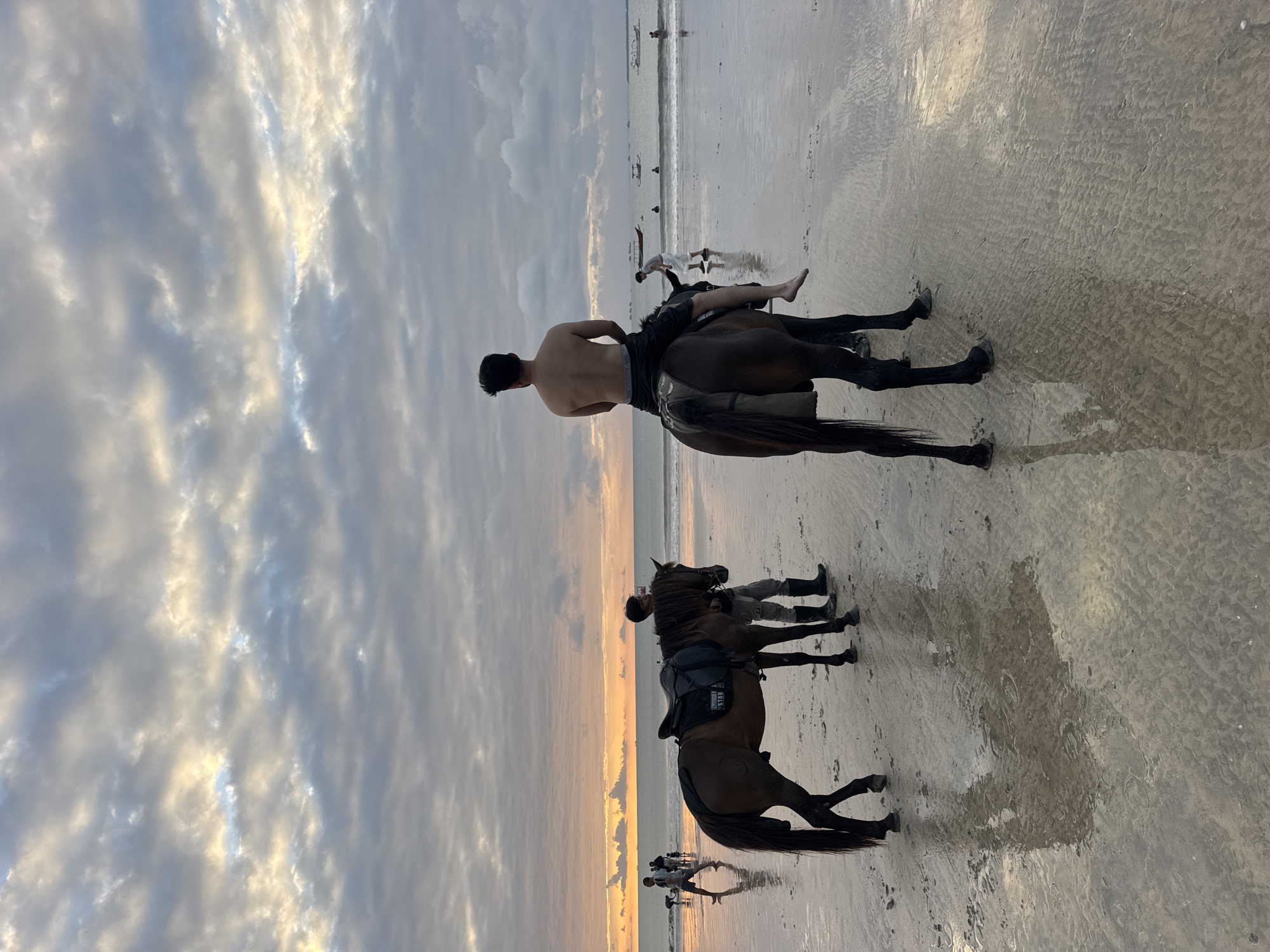 This media file depicts a group of people on a beach in Kuta Selatan, Indonesia, at sunset. The primary subjects are several individuals, including some shirtless men, riding horses on the wet sand. The setting is a tranquil beach with calm waves and reflections on the wet sand. The sky is filled with dramatic clouds, illuminated by the warm hues of the setting sun, suggesting a serene and beautiful evening.
The activity taking place is horseback riding on the beach. The people appear to be enjoying a leisurely ride along the shoreline. One rider, a shirtless man, is positioned in the foreground, facing away from the camera. Another horse and rider are visible in the middle ground, with a third horse further back. Several other figures are also present on the beach, some in the water and others walking.
Notable details include the overcast yet colorful sky, indicative of sunset. The wet sand creates reflections of the sky and the horses, adding depth and visual interest to the scene. The presence of boats in the distance suggests proximity to the sea. The overall mood of the image is peaceful and relaxed, capturing a beautiful moment at the end of the day. No visible text is present in the image.