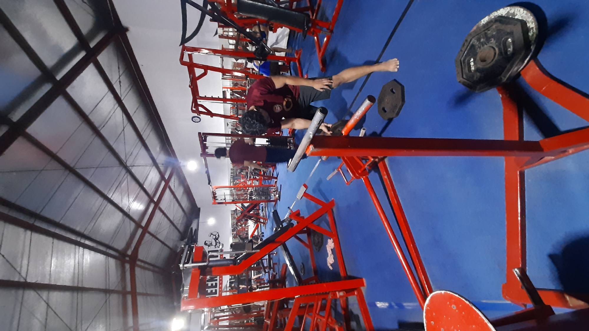 This image shows the interior of a gym located in Sedayu, Indonesia. The gym features a blue mat floor and a metal ceiling with visible beams and lights. Red exercise equipment, including weight machines and benches, is arranged throughout the space. In the foreground, a young man wearing a maroon t-shirt and black shorts is standing and appears to be interacting with weight plates on the floor. Another man, also in a maroon t-shirt, is visible in the middle ground, walking away from the camera. Several other individuals are present in the background, engaged in various gym activities, with mirrors lining the back wall. The overall impression is that of a functional and active fitness facility.