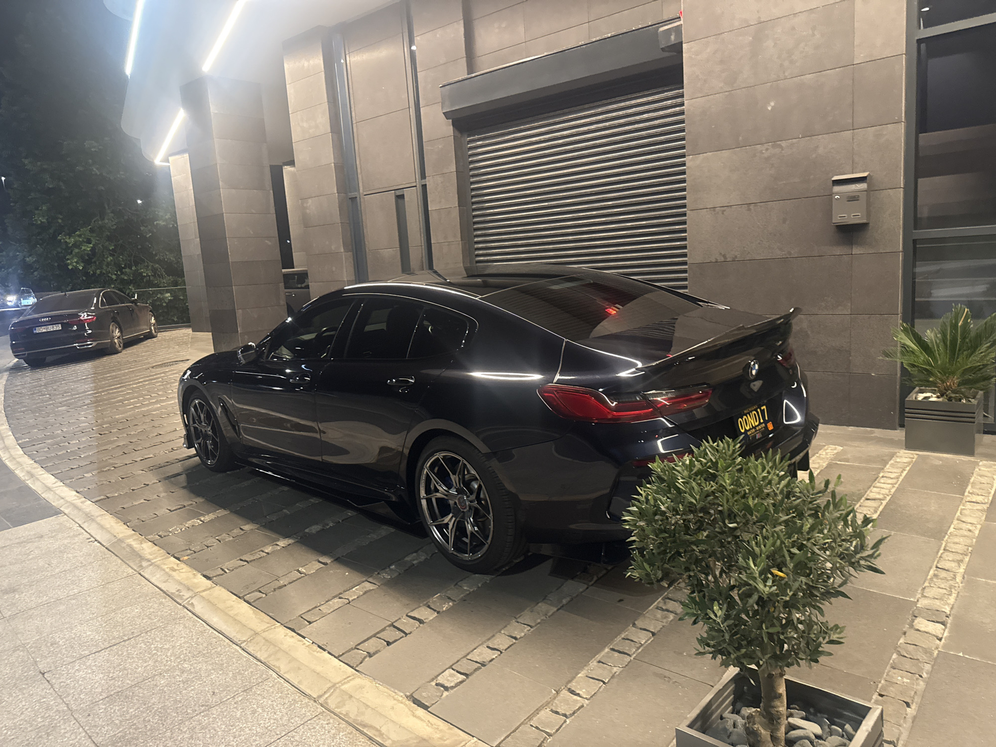 A black BMW sedan with a license plate reading "OONDI17" is parked on a stone-paved driveway in front of a modern building with large pillars and a corrugated metal garage door. The car's rear is visible, and a small green tree in a pot is positioned to the right of the vehicle. The scene appears to be at night, with artificial lighting illuminating the area. Another black car is visible in the distance on the left side of the image.