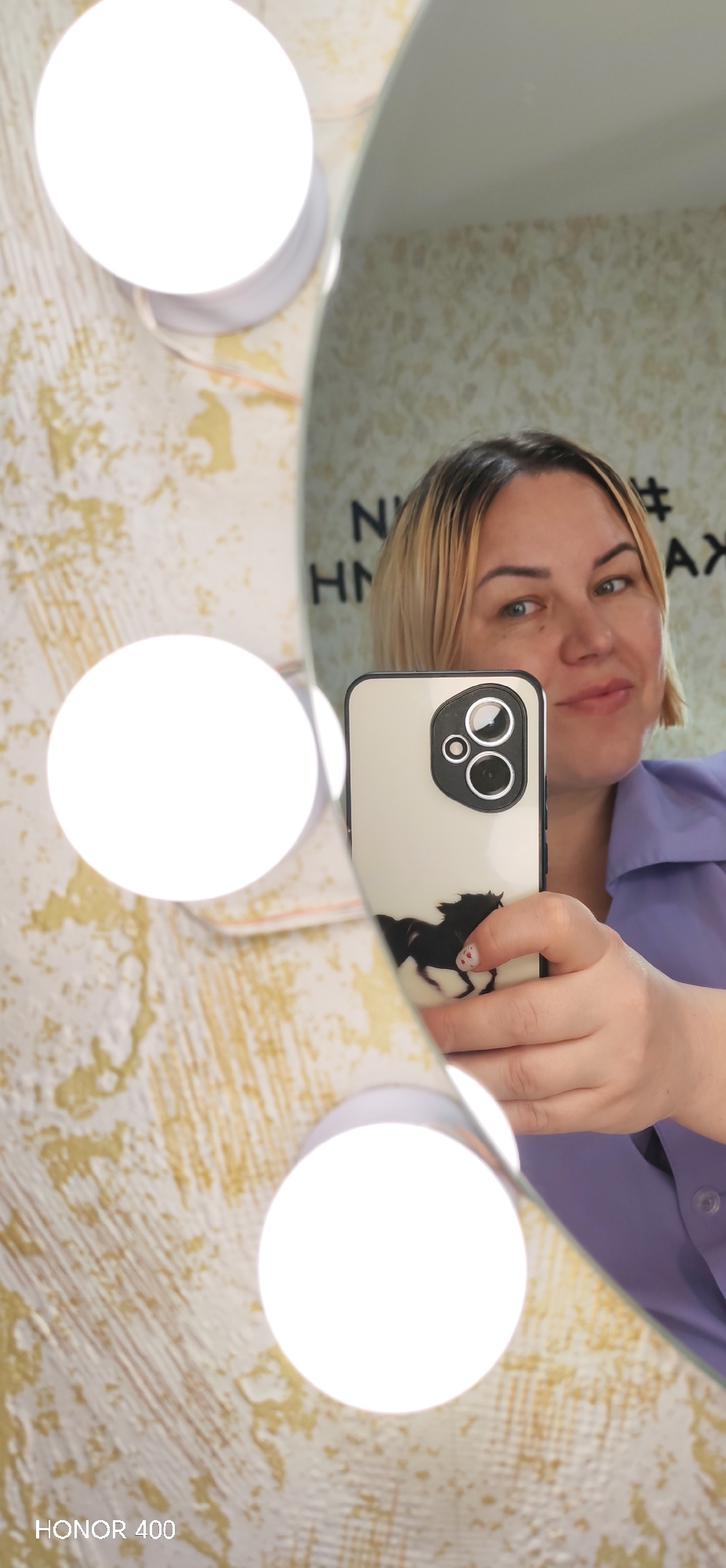 A mirror selfie captures a woman holding a light-colored smartphone, which is also reflected in the mirror. The woman has shoulder-length blonde hair with darker roots and light-colored eyes, and she is wearing a light purple collared shirt. She has a subtle smile on her face. Her right hand, holding the phone, shows manicured nails, with a small red heart design visible on her pinky finger.
The smartphone case is light-colored and features a distinct black silhouette of a horse on its lower half. The phone's camera module, visible in black, contains multiple lenses. To the left of the mirror's reflection, three large, round, bright white vanity lights are prominently featured, contributing to the well-lit scene.
The background visible in the mirror reflection is a light-colored wall with a textured or patterned surface, possibly wallpaper in beige and light brown tones. Faint text is visible on this wall, including "N #" and "HMA AX". A watermark "HONOR 400" is present in the bottom left corner of the image, indicating the device that captured the photo. The setting appears to be indoors, likely a personal space with a vanity or dressing mirror.