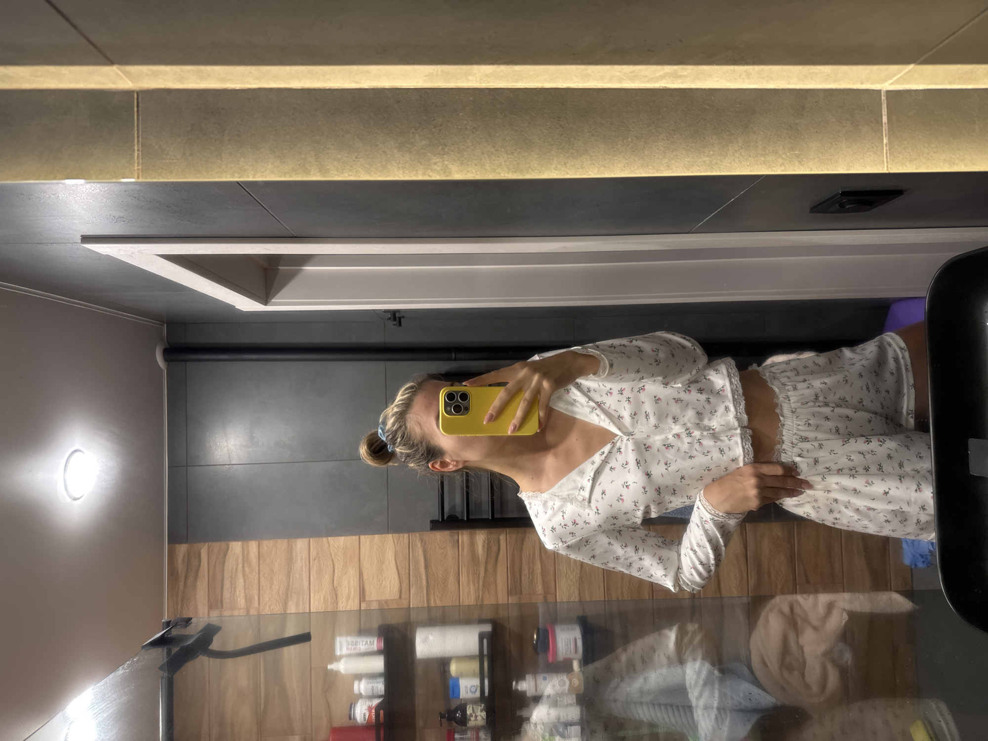 A woman is taking a selfie in a bathroom. She is wearing a white floral print pajama set. The bathroom has gray tile walls and wood paneling, with a lit mirror above a vanity. The vanity has items stored on shelves and a black countertop.
