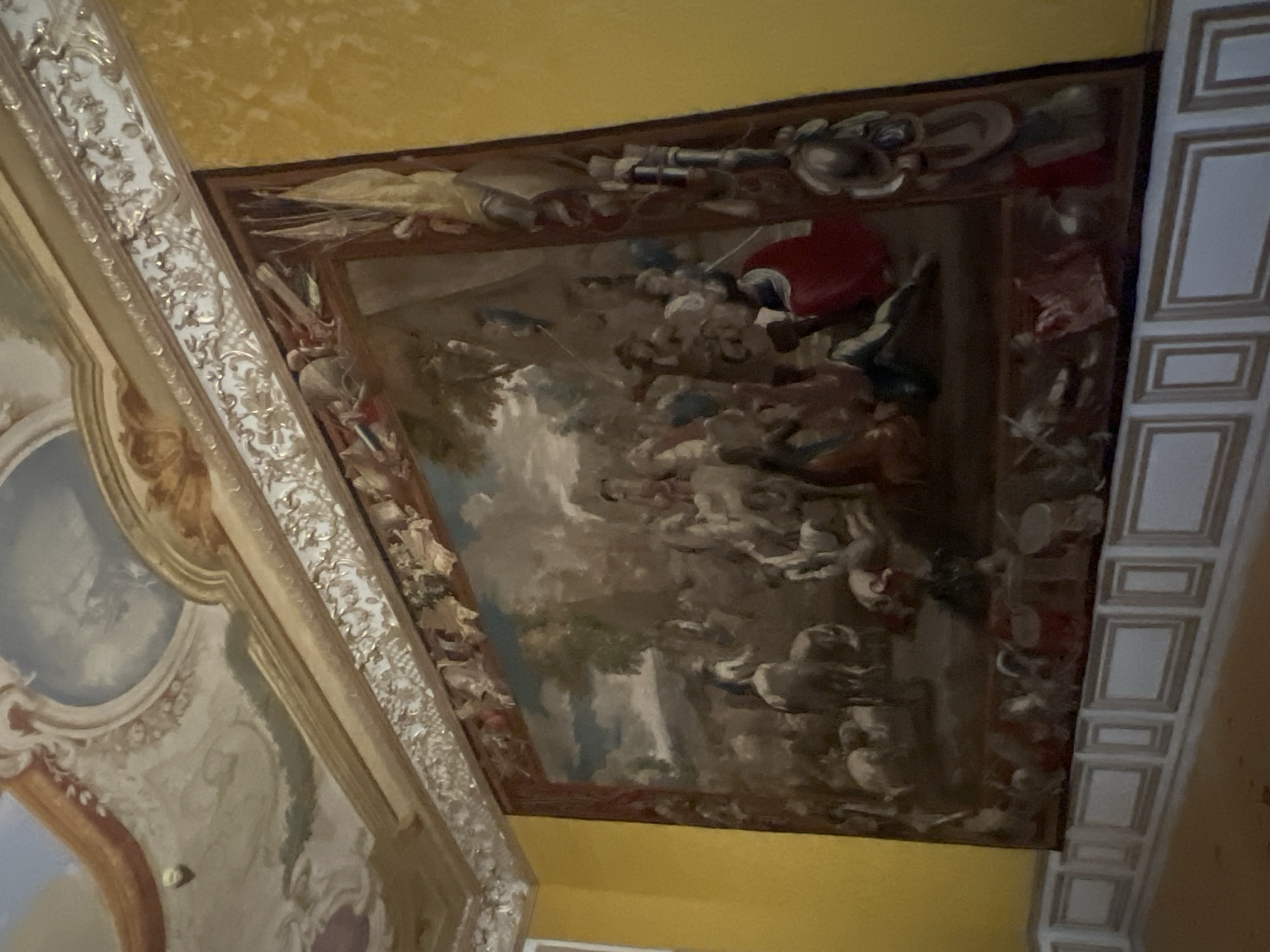 The image shows a tapestry hanging on a yellow wall in a room with ornate ceiling decorations. The tapestry depicts a busy scene with many figures, some on horseback, and animals. The style of the tapestry suggests it is a historical artwork. This is likely the New Schleissheim Palace in Oberschleissheim, Germany, which is known for its opulent interiors and extensive art collection. The room appears to be a reception or grand hall, typical of a palace setting. The perspective of the photo is from a low angle, looking up towards the tapestry and the ceiling.