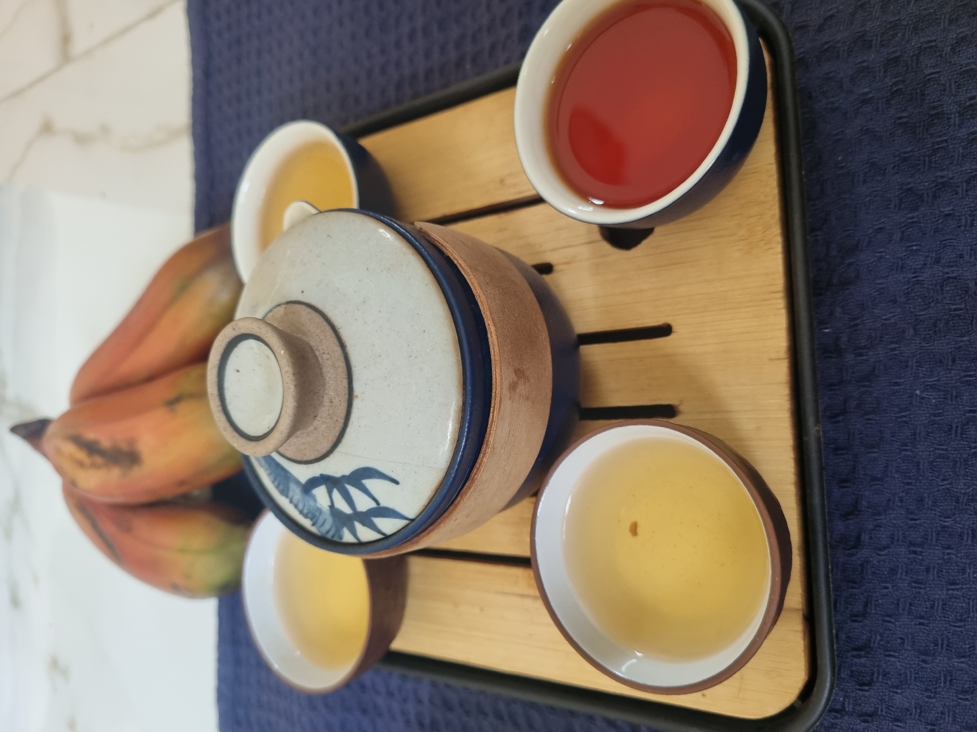 On a wooden tray atop a blue textured surface lies a tea set consisting of a teapot and four teacups. The teapot is decorated with a white lid bearing blue floral patterns. Two teacups contain a light-colored liquid, while another holds a dark red liquid. A group of reddish-orange bananas sits next to the tea set. The setting is undetermined, and there is no indication of the location being Pedda Kammavaripalli, India. There are no people in the frame, and no actions or interactions are observable.