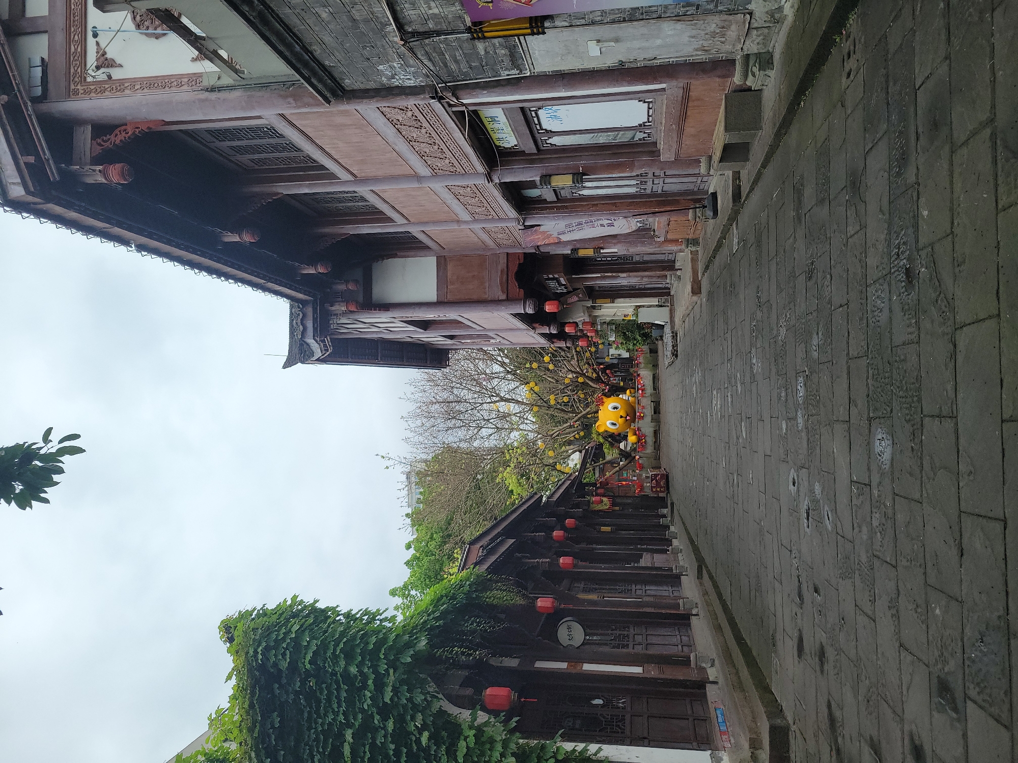 The image shows a street in Hechuan, China. The street is paved with gray stone bricks. Buildings line both sides of the street, featuring traditional Chinese architecture with wooden frames and intricate carvings. Some buildings are adorned with red lanterns. Lush greenery, including climbing vines, covers parts of some buildings. At the end of the street there is a yellow tiger display. The sky is overcast.