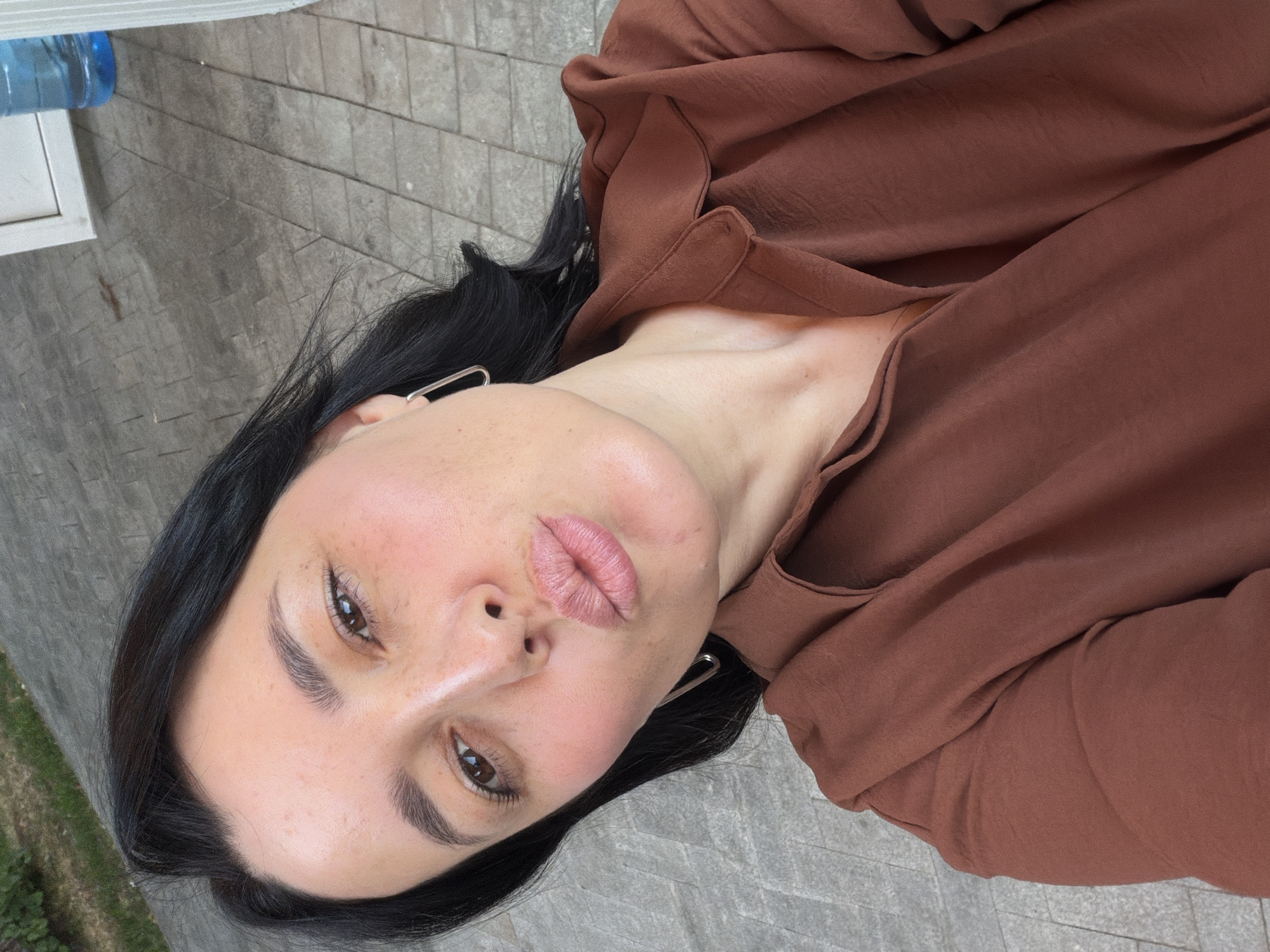 A selfie of a woman with dark hair and light skin, wearing a brown shirt, is taken outdoors. The woman is puckering her lips, and appears to be wearing subtle makeup, including blush and lip gloss. She has light brown eyes and a few freckles on her cheeks. She is wearing a silver, geometric earring in her visible ear. In the background, a tiled or paved area is visible, as is some green foliage. To the right, a blue plastic bottle of water is visible on a white surface. The lighting suggests it is daytime. The provided information about Moscow, Russia is not supported by visual evidence in the image.