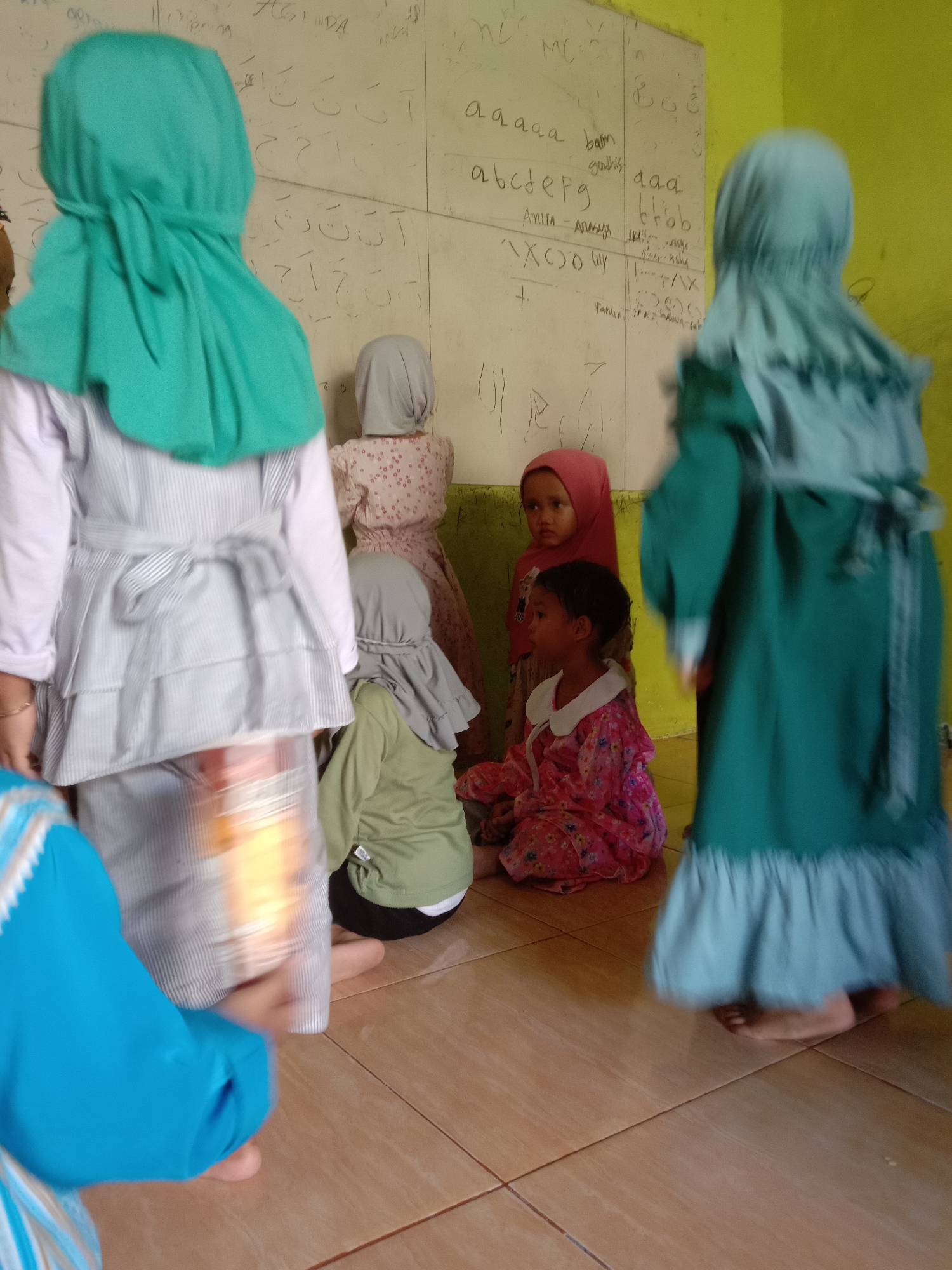 In a classroom in Tulungrejo, Indonesia, several young girls wearing hijabs stand and sit near a whiteboard covered with Arabic script and English letters. One girl in a turquoise hijab is facing away from the camera, her back to the whiteboard, wearing a striped grey dress. To her left, another girl in a grey hijab, also facing away, wears a floral dress. In front of them, a girl in a pink hijab is seated, looking to her left, while another child in a pink floral dress sits beside her. Another girl in a light green hijab is seated on the floor, her back to the camera. A child in a bright blue and white patterned garment is partially visible on the left, with their legs crossed. The room has yellow walls and a tiled floor. The whiteboard contains handwritten Arabic characters, the English alphabet (e.g., "abcdefg", "aaaaa"), and various other markings.