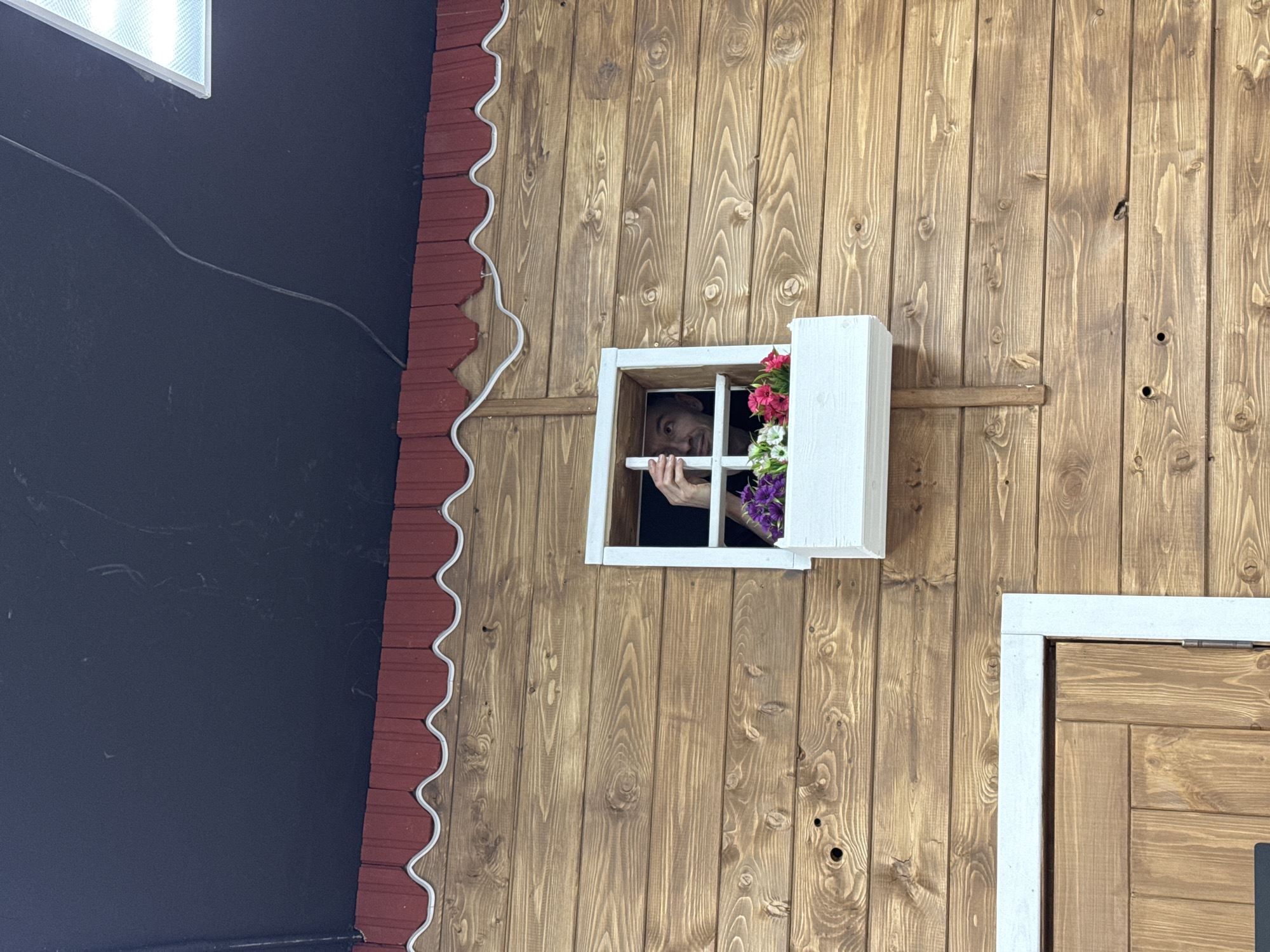 A medium shot of a human peering through a window. The setting appears to be an inverted or upside-down room exhibit, possibly for photo opportunities. The room features wooden plank walls and a white-framed window with flowers. The person is male with dark hair, and he is looking out with a playful expression. Part of a black wall with a faux roof is also visible. The location is identified as Izhevsk, Russia.
