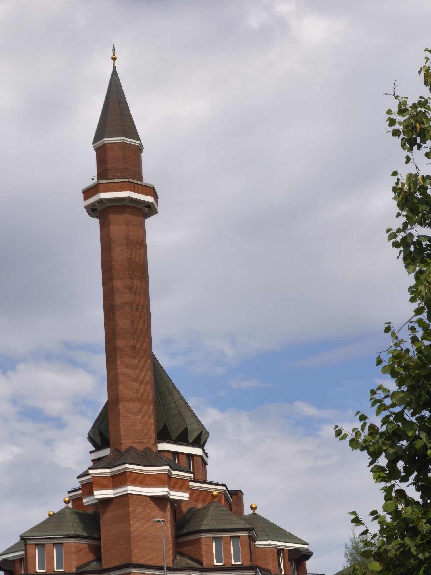 A close-up, low-angle shot captures the upper portion of a red brick minaret and adjacent structures against a cloudy sky. The minaret, octagonal in shape, tapers towards a pointed, dark roof topped with a small gold spire. White trim decorates the base of the upper section of the minaret. Below the minaret, several green-tiled roofs with white trim and gold finials are visible. A cluster of green leaves and branches from a tree occupies the right side of the frame, partially obscuring the sky. The overall scene is devoid of people and any discernible signs of human activity. The location is identified as Unknown, Unknown.