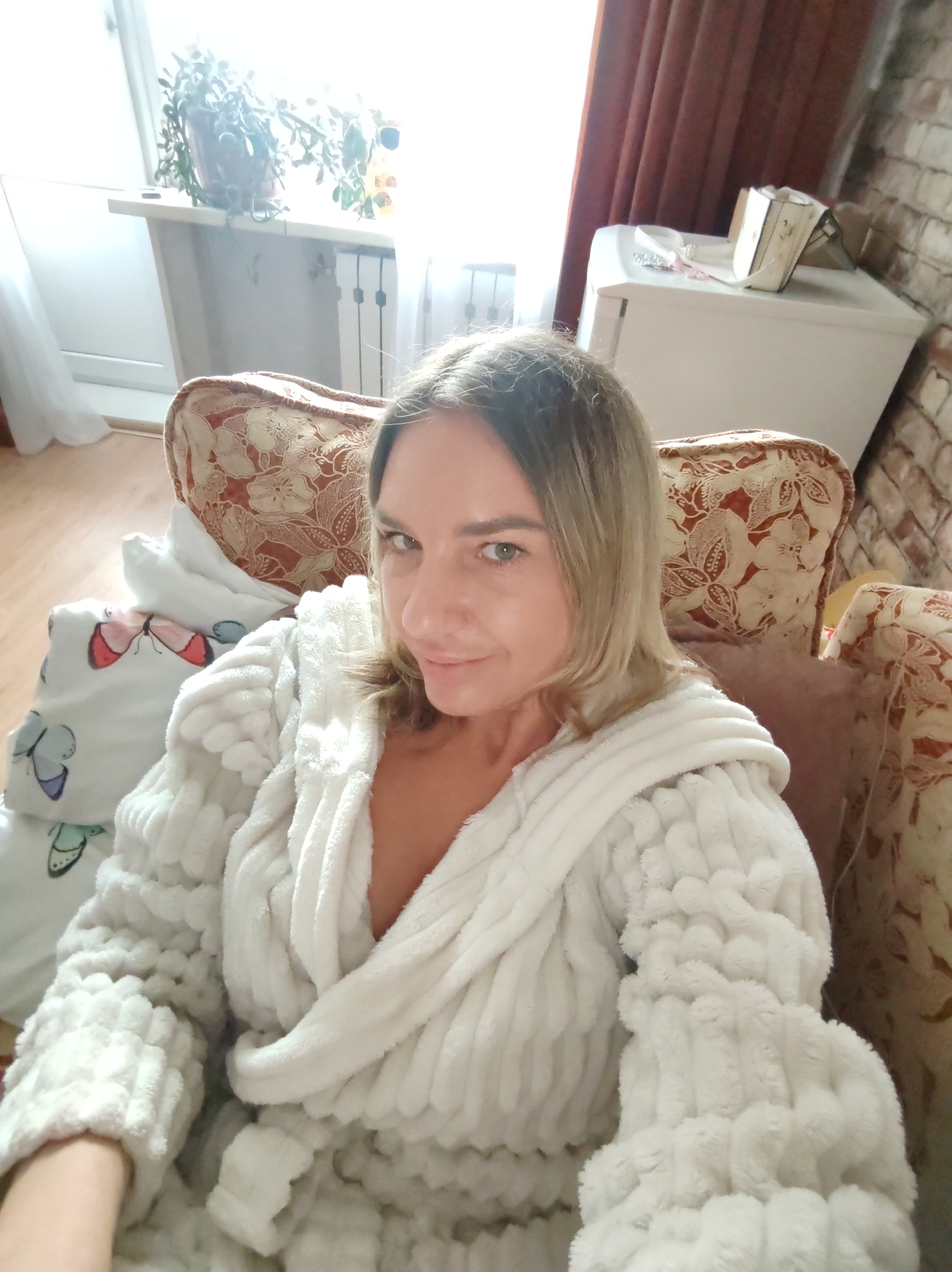 The photograph presents a woman in a white, plush robe, seated on a floral patterned armchair. Her hair is blonde and shoulder-length. She is looking directly at the camera with a slight smile.
Behind her, there is a window with a potted plant on the sill and a white cabinet. The room has a brick wall. The background indicates an interior setting, but there are no specifics in the image to identify a location in Petropavlovsk-Kamchatsky, Russia.