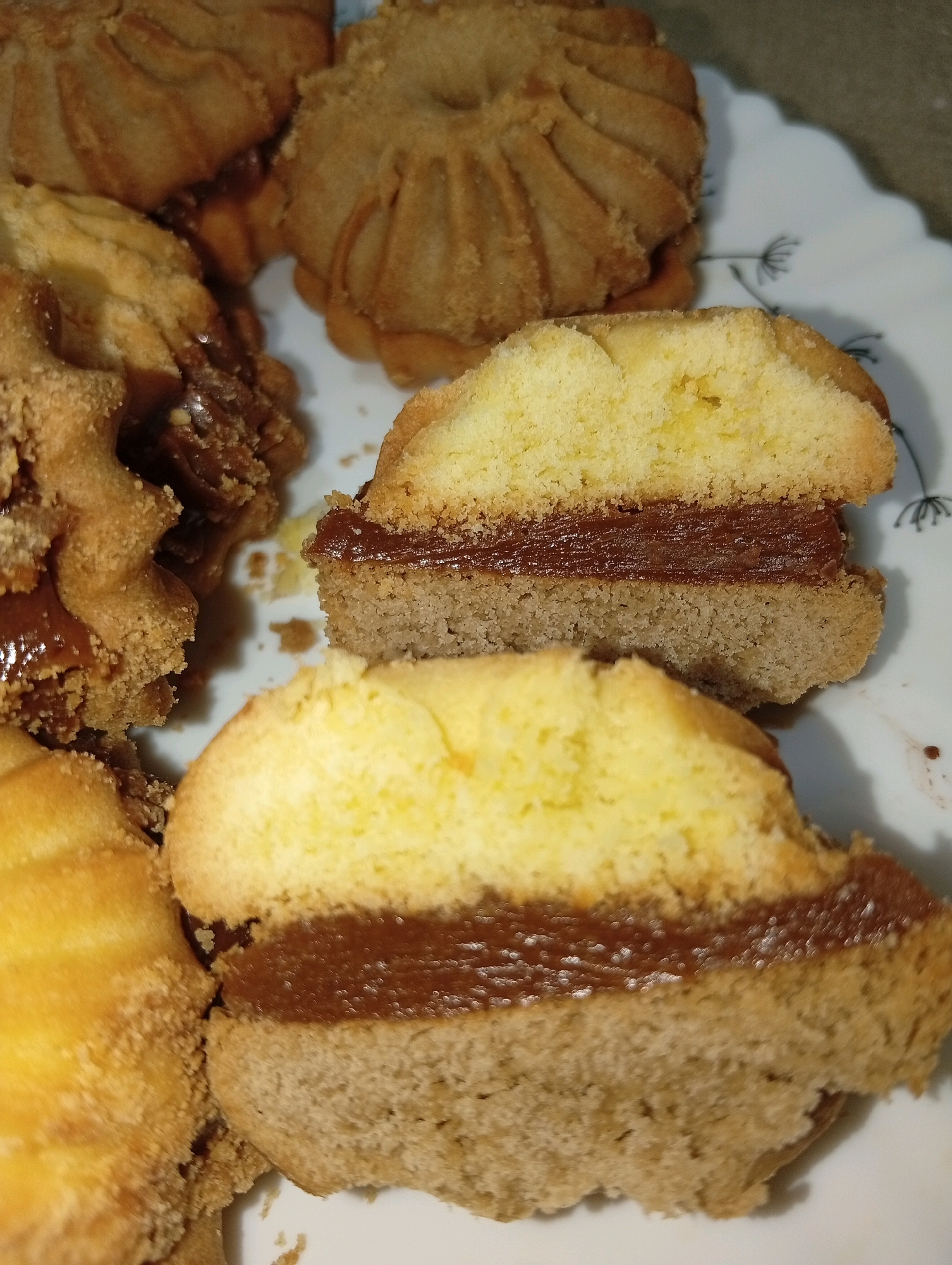 A close-up, overhead shot shows a plate of alfajores, which are sandwich cookies with a dulce de leche filling. The cookies are arranged on a white plate with a faint floral pattern. In the foreground, two alfajores are visible, one of which has been cut in half, revealing the light yellow cake and dark brown dulce de leche filling. The cookies have a golden-brown color and a slightly crumbly texture. The background shows more intact alfajores, with their ridged edges and similar filling. The lighting is warm and natural, highlighting the textures and colors of the baked goods.