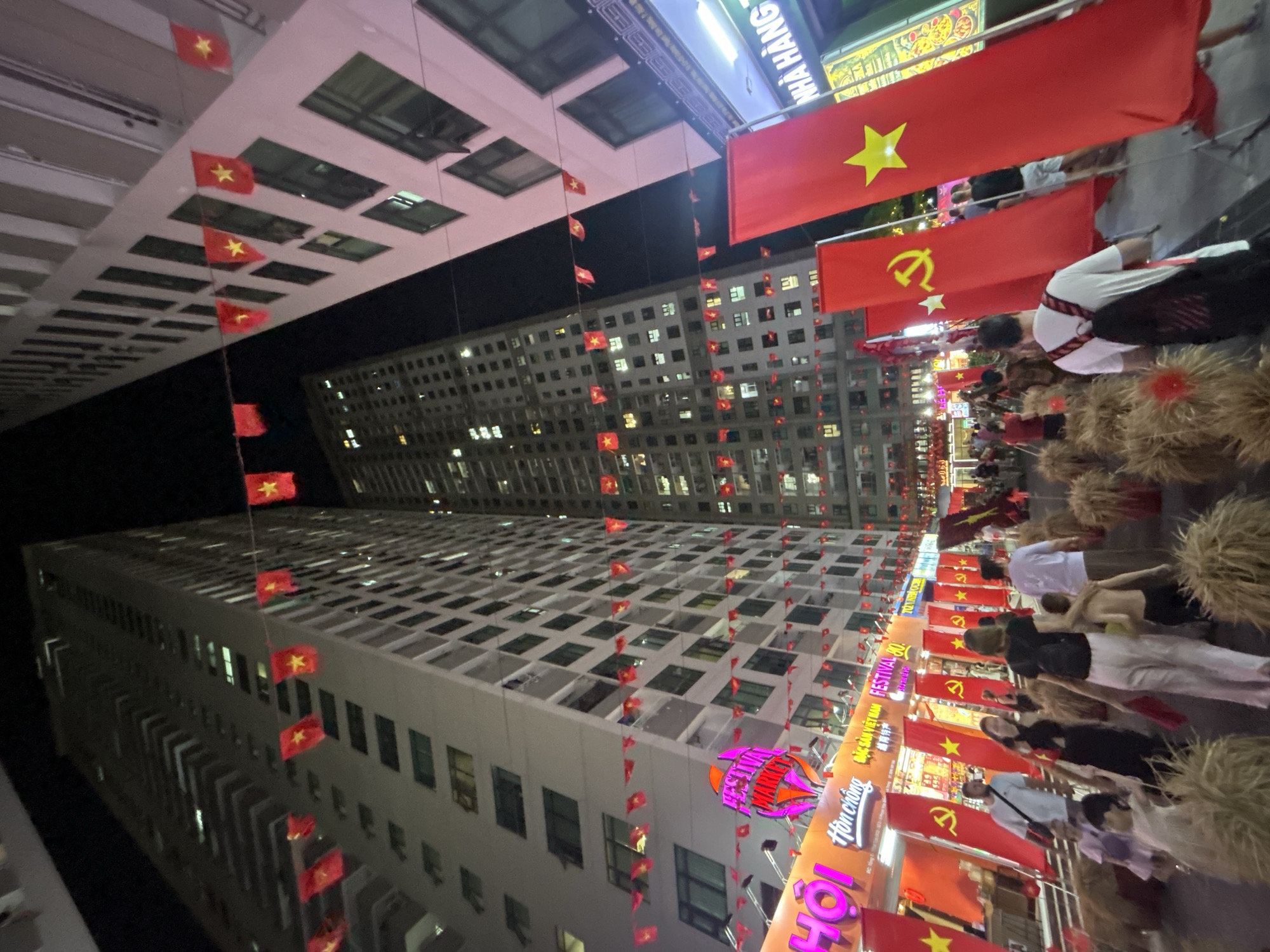 A night view in Bắc Nha Trang, Vietnam, features high-rise buildings decorated with Vietnamese flags. Strings of small flags run vertically between the buildings. Large red flags with yellow stars and communist symbols hang above a street crowded with people. Some individuals wear traditional clothing and carry bundles. The scene is illuminated by streetlights and storefronts. Signage with Vietnamese writing is visible.
