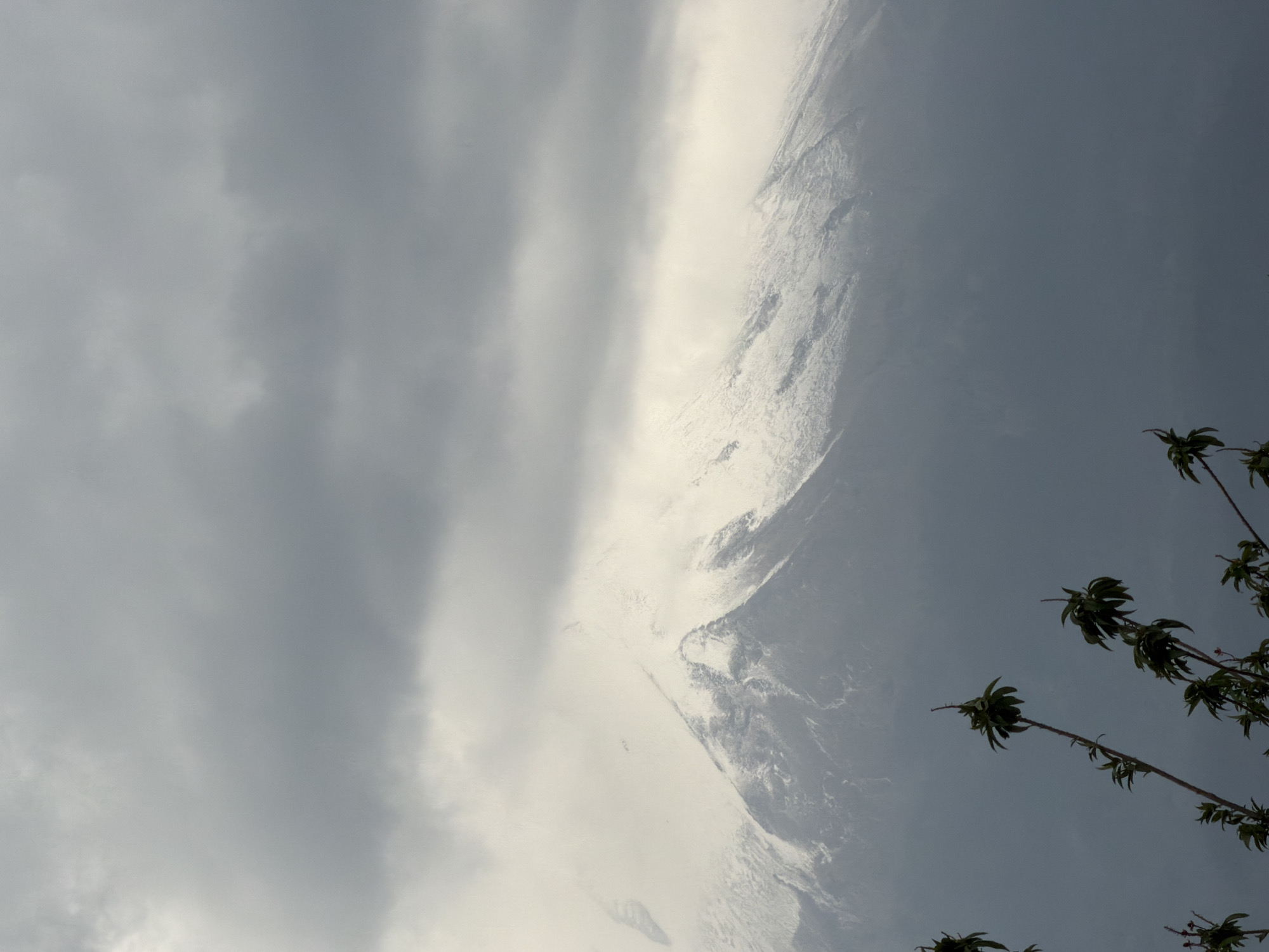 A low-angle shot shows a partly cloudy sky over Metepec, Mexico, with the snow-covered peak of the Popocatépetl volcano visible through a break in the clouds. Some foliage is visible in the foreground.
