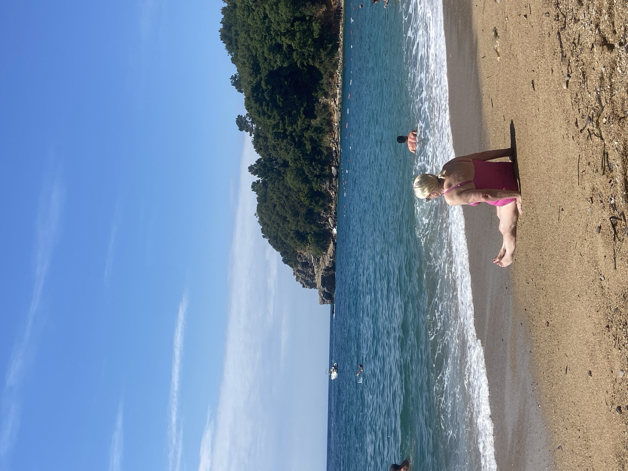 An outdoor, eye-level shot captures a vibrant coastal scene, likely in Sveti Stefan, Montenegro, on a bright, sunny day. The image features a sandy beach meeting clear, blue-green sea, bordered by a lush, green, forested hillside or cliff.
In the foreground, a woman with blonde hair, wearing a pink one-piece swimsuit, stands near the water's edge with her back partially turned to the viewer. Her shadow is cast behind her on the wet sand. Gentle white-capped waves lap at the shore around her feet.
Several other people are visible in the mid-ground, swimming or wading in the sea. One person's upper body is seen closer to the shore, while others appear further out. A line of white and red buoys stretches across the water, likely delineating a swimming area. In the distance, a small white boat or yacht is anchored or moving across the water.
The background is dominated by a dense, dark green forest covering a steep landmass that plunges into the sea. The sky above is a clear, bright blue with a few wispy white clouds, indicating excellent weather. The overall impression is one of leisure and natural beauty, typical of a summer day at the beach. There is no visible text.