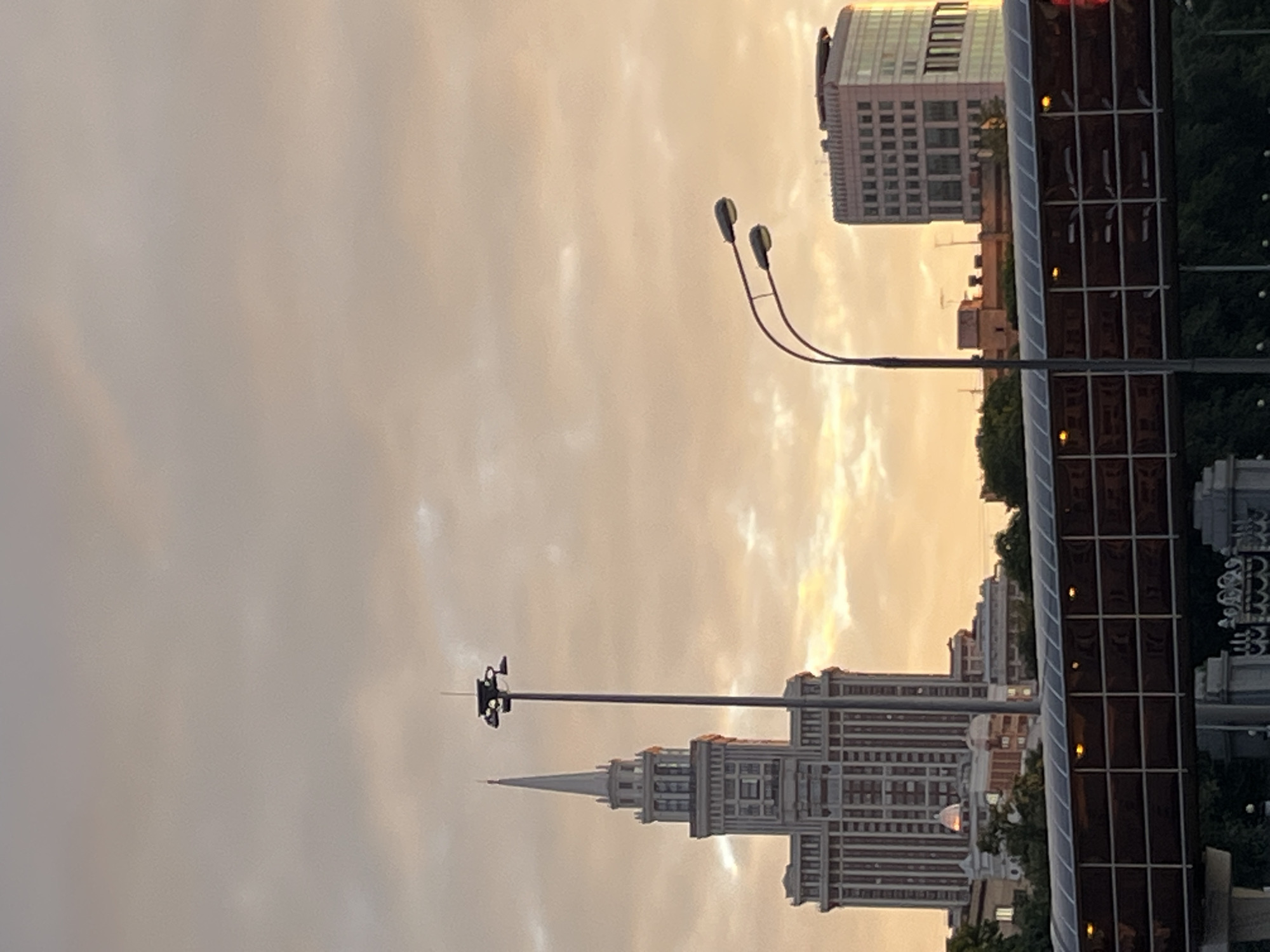 In Khimki, Russia, a panoramic view captures a cityscape at dusk. The sky, a canvas of soft, warm hues, is filled with scattered clouds tinged with the golden light of the setting sun. Dominating the left side of the frame is a tall, art deco-style skyscraper, its intricate facade and pointed spire reaching towards the heavens. Beside it, a tall street lamp with multiple lights stands sentinel. In the mid-ground, a modern, rectangular building with a glass facade reflects the twilight sky. In the foreground, a glass-enclosed pedestrian bridge spans across the scene, its internal lights casting a warm glow. The bridge is supported by concrete structures. The base of the bridge is lined with a decorative metal fence. Lush green trees are visible behind the bridge, adding a touch of nature to the urban landscape. The overall impression is one of serene urban beauty as day transitions into night.