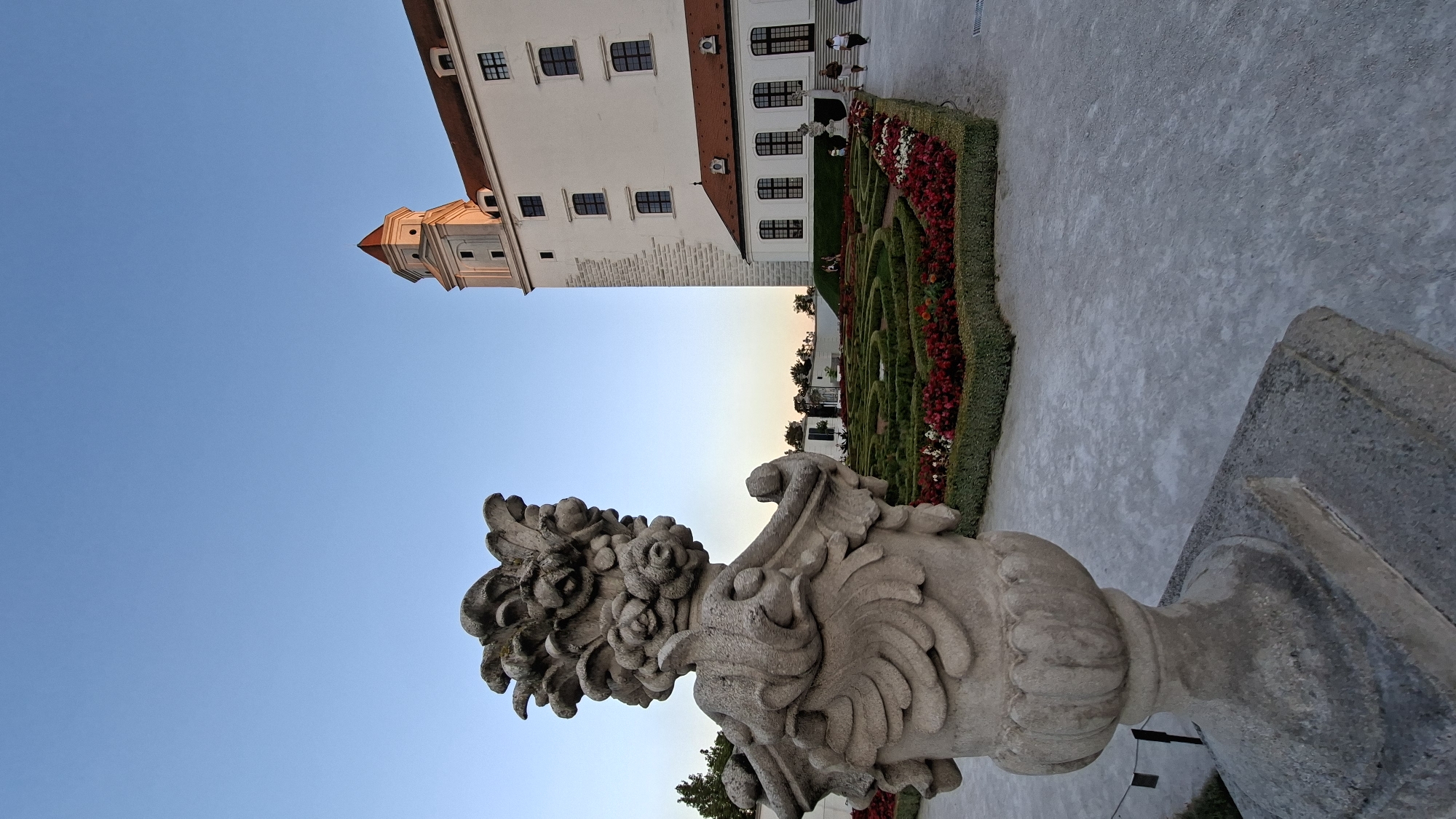 The image depicts Bratislava Castle in Bratislava, Slovakia, during a clear day with a bright blue sky. In the foreground, a large, ornate stone urn with intricate floral carvings stands to the left, positioned on a square stone base. Behind the urn, a meticulously maintained formal garden is visible, featuring geometric hedges and vibrant red and white flowers. Beyond the garden, the white facade of Bratislava Castle rises, with its distinctive reddish-brown tiled roof and a tall, white tower topped with a similar roof and red accents. Several windows are visible on the castle walls. A few people are casually walking on the paved pathway to the right of the garden, suggesting a public space. The overall scene conveys a sense of historical grandeur and well-kept landscape.