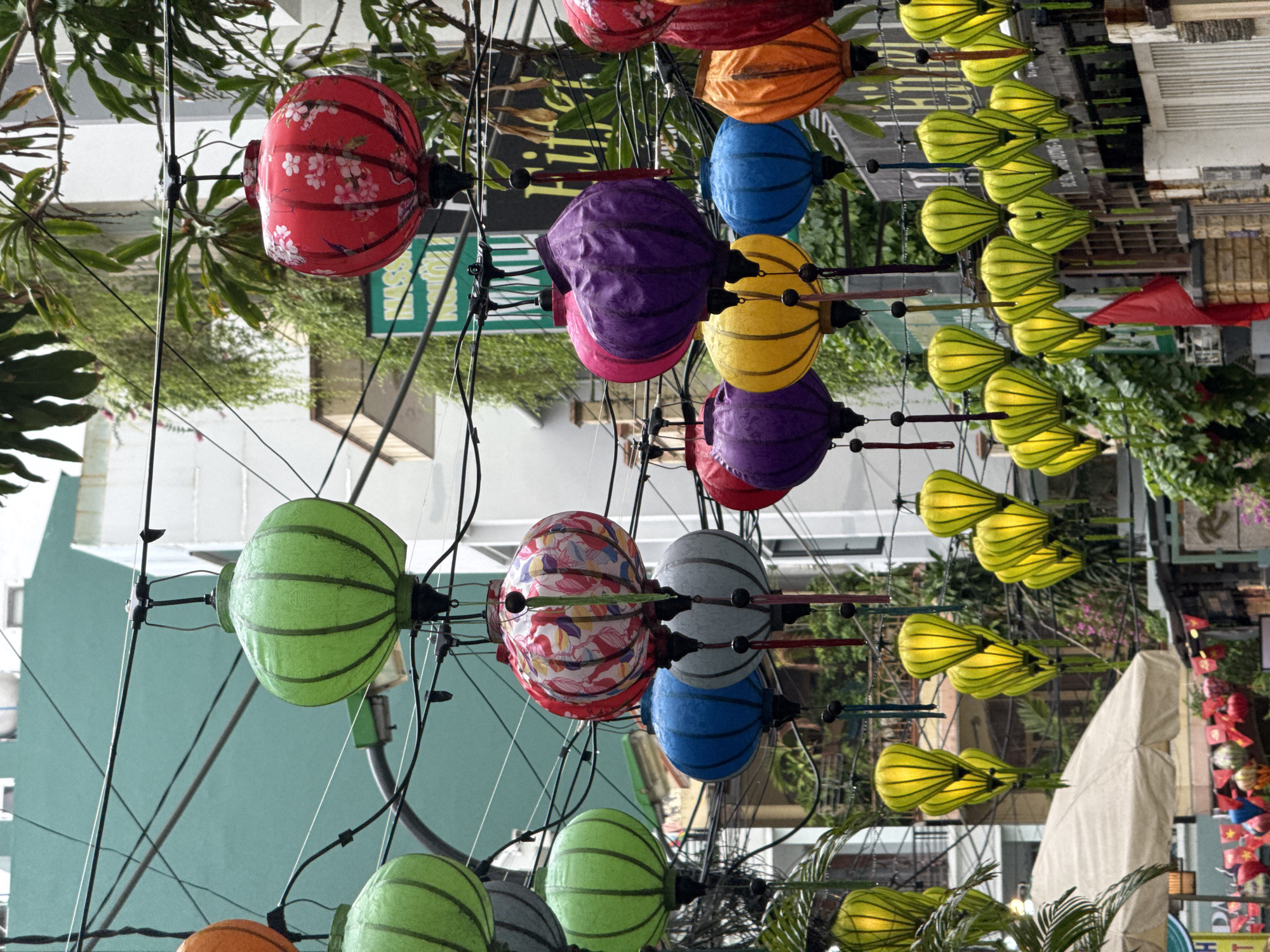 The image shows a street scene in Nha Trang Ward, Vietnam, decorated with colorful lanterns. Several strings of lanterns are hanging overhead, including red, purple, yellow, blue, green, and gray lanterns. Some have patterns, like flowers or stripes. Thin wires support the lanterns. Buildings and greenery are visible in the background. A white canvas tent and red Vietnamese flags are seen near a shop. The lanterns create a festive atmosphere.