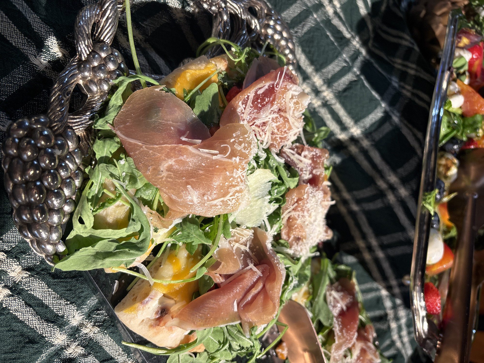 This is a close-up, high-angle, eye-level shot of two plates of food on a green and white checkered tablecloth. The plate in the foreground, a circular silver platter with a decorative grape cluster design on the left, contains pieces of pizza topped with prosciutto, arugula, and grated cheese. The pizza appears to have a yellow sauce base. Another plate of food, partially visible in the background, is a silver rectangular tray with more food that includes red berries, blueberries, and what appears to be mozzarella cheese and fruit. The lighting suggests it is daytime, with sunlight casting shadows. The setting is an indoor or outdoor dining area, possibly a restaurant or a catered event. There is no specific indication of the location being in Riga, Latvia.