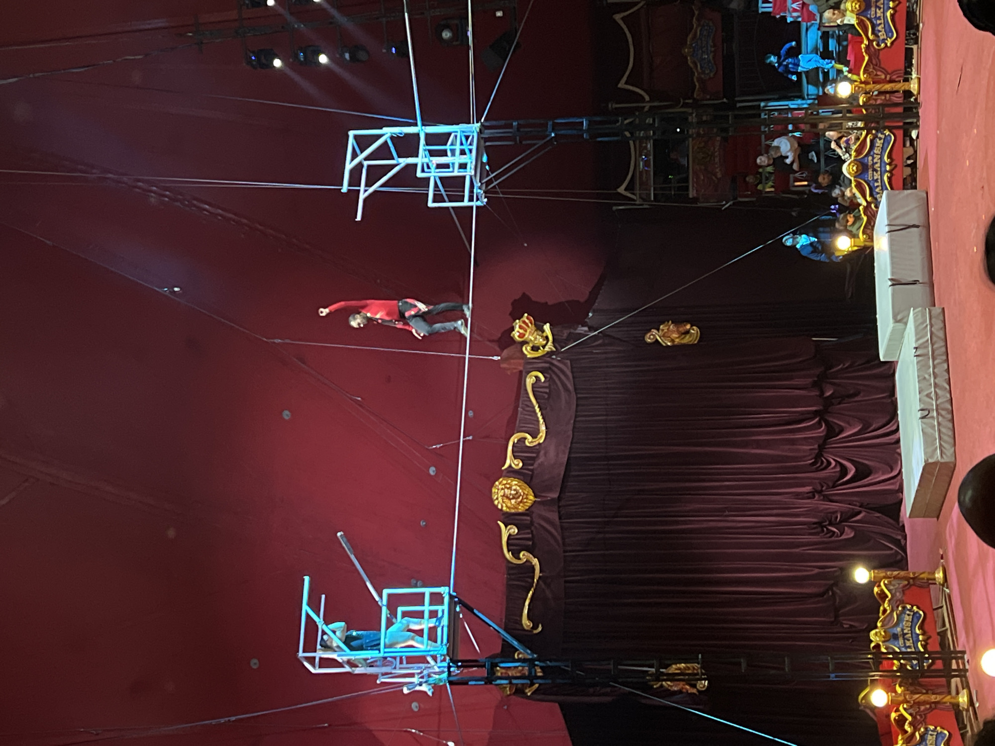 The image depicts a circus performance. A male acrobat wearing a red and black outfit is seen lying prone on a tightrope, spanning between two platforms. On one platform, another individual is lying on their back, possibly preparing for an act. In the background, theater curtains and stage elements can be observed. Audience members are visible in the upper background. A partial sign reading "BALKANSKI" suggests a connection to the Balkans region, although the exact city and location are unknown.