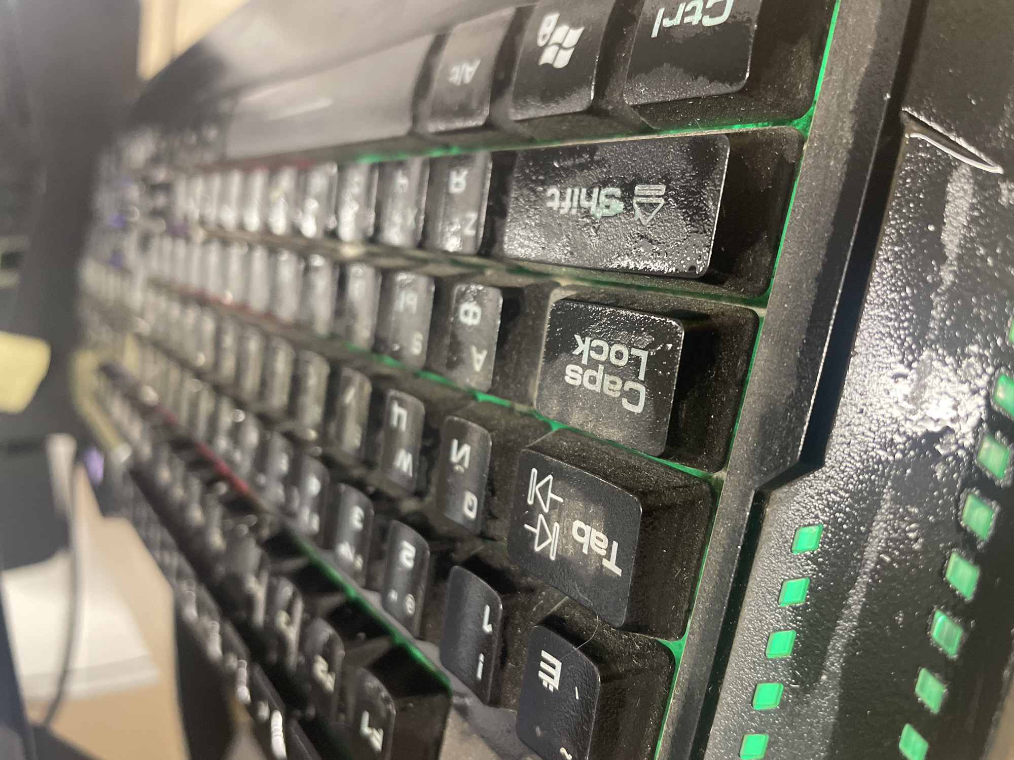 The image shows a close-up, low-angle view of a dirty, black computer keyboard with green backlighting. Several keys are visible, including "Tab," "Caps Lock," "Shift," and "Ctrl." There are also keys with what appear to be Cyrillic characters and numbers. Dust is visible on the keys and the surrounding keyboard surface. The lighting suggests an indoor environment, possibly an office or home setting. There are no people or discernable city-related features present in the image. The location of Терволово, Russia, is not visually represented in this photograph.