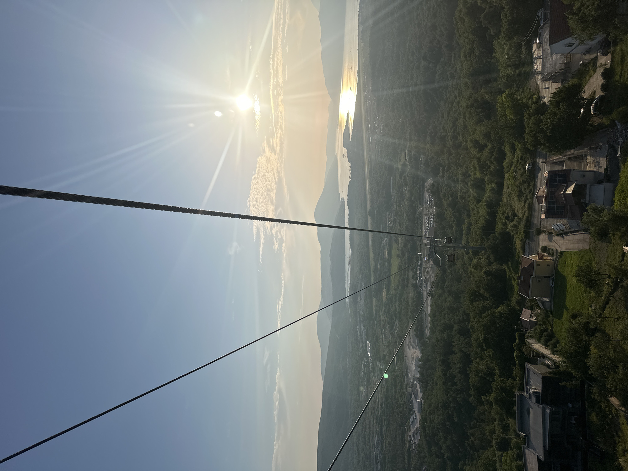 The image captures a scenic view from a cable car ascending towards the peak of Dub, Montenegro, overlooking a bay and lush green hills. The sun, positioned high in the sky, casts a warm, golden glow across the landscape, creating lens flares and highlighting the wispy clouds. In the foreground, the thick, dark cable of the cable car stretches diagonally across the frame, with a single cabin visible in the distance, also suspended from the cable.
Below, the terrain transitions from a cluster of houses with red-tiled roofs and solar panels in the immediate foreground to a dense forest that blankets the slopes. Further in the distance, the bay unfolds, dotted with small boats and reflecting the sunlight. Mountains form a majestic backdrop to the bay, their forms softened by the haze of the late afternoon sun.
The overall impression is one of tranquility and natural beauty, amplified by the elevated perspective offered by the cable car. The scene depicts a typical day in the summer, with clear skies and pleasant weather, ideal for outdoor activities and sightseeing. The presence of buildings and roads suggests human habitation and infrastructure integrated into the natural landscape. The cable car itself signifies a tourist attraction or a mode of transportation designed to provide visitors with breathtaking panoramic views of the region.