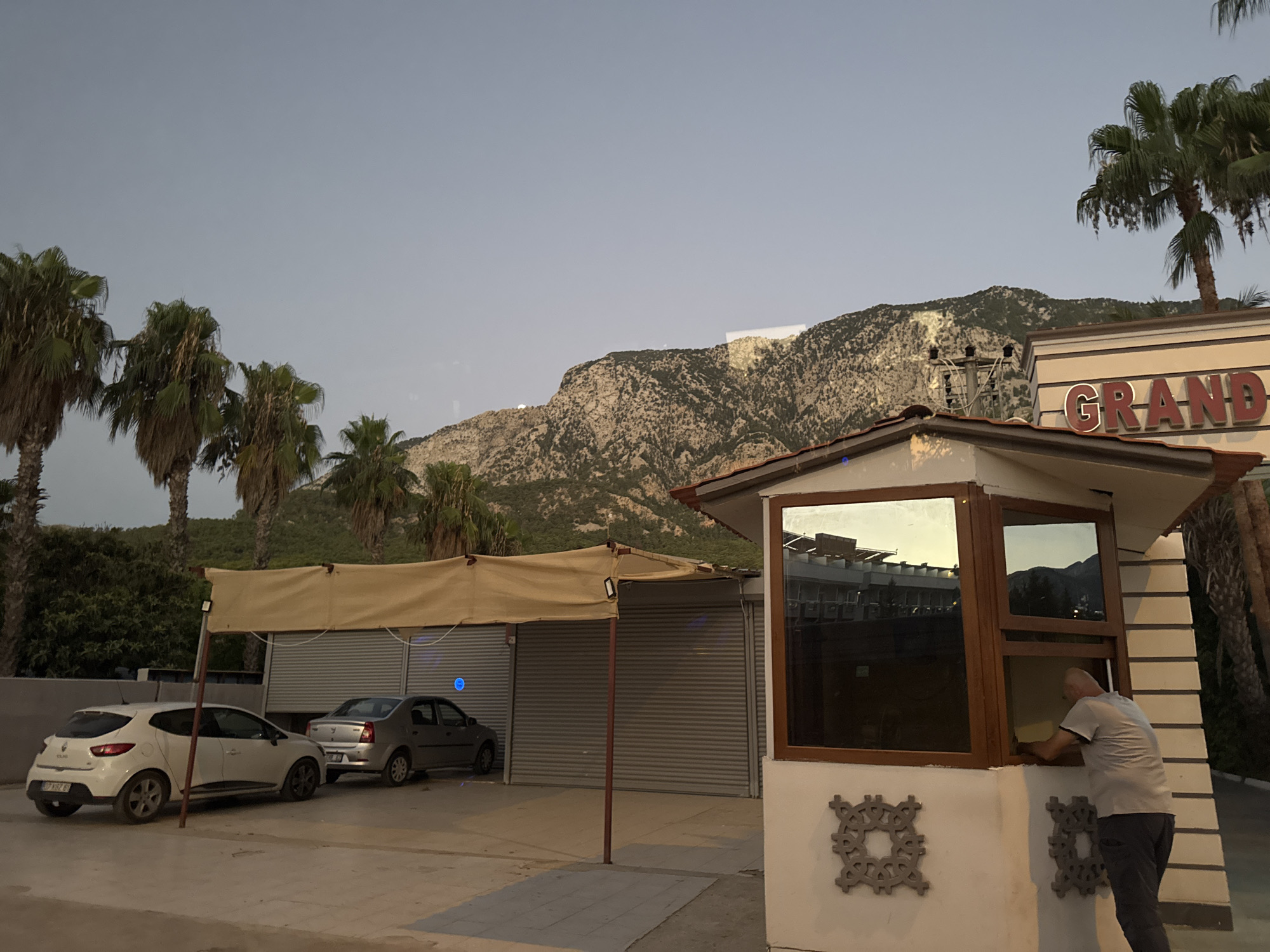 A man in a gray t-shirt and dark pants is leaning out of a window of a small white building. The building has a brown trim around the window and decorative gray medallions on the wall. Behind the building, palm trees line a mountain in the background, with a clear sky above. In front of the building, a white car and a silver car are parked. A tan awning stretches over a row of gray, corrugated metal doors. The scene appears to be set in the city of Kemer, Turkey, as suggested by the typical Mediterranean flora and architecture.